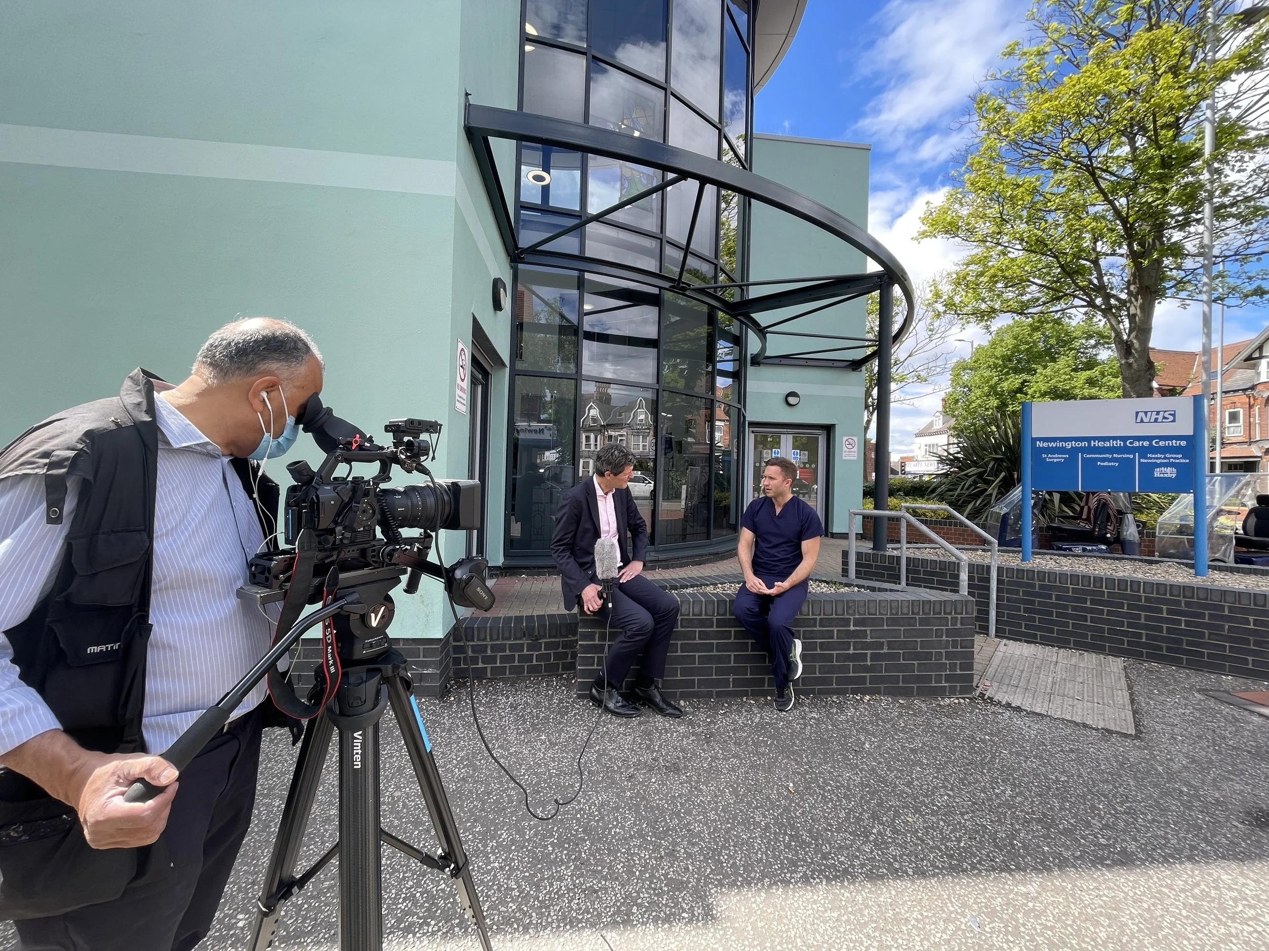 BBC Chief Health Editor Hugh Pym holding a microphone while interviewing Dr. Mathew Fortnam outside of a GP practice in Hull.