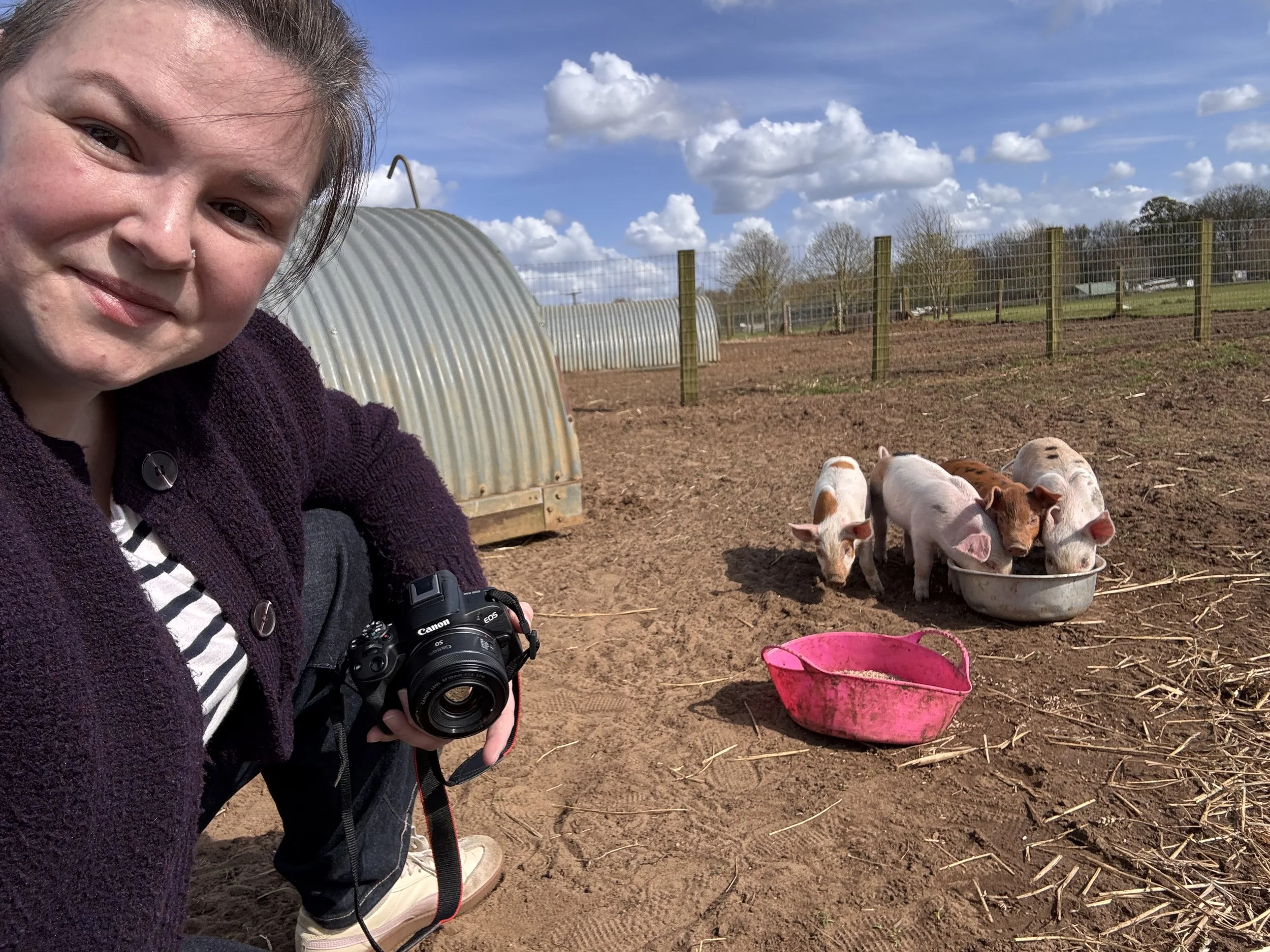 Melissa Cooper holding a camera takes a selfie on a farm during a Content Day. In the background, piglets are gathered near a pink bucket and a feeding trough. The scene includes a fenced area with trees and a partly cloudy sky.