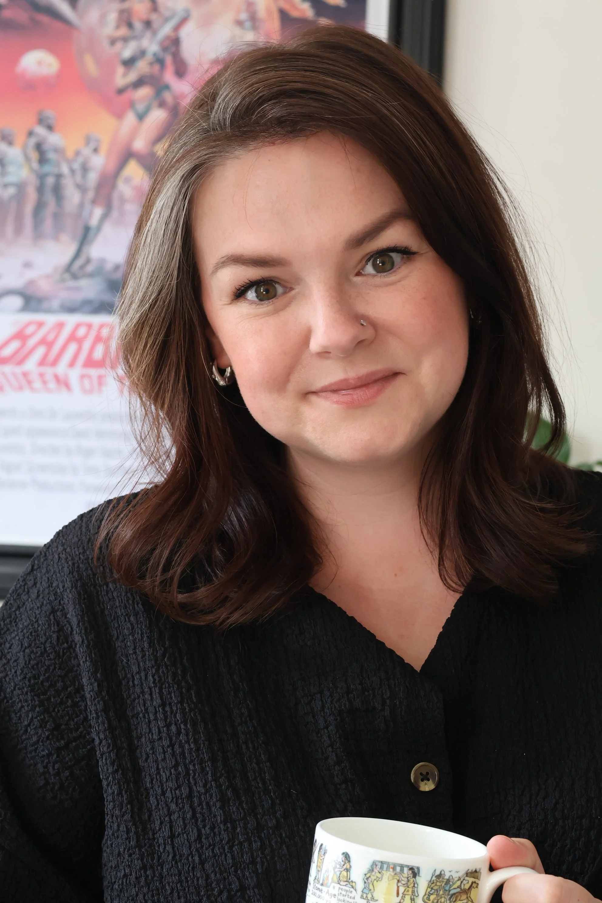 Melissa Cooper, a woman with medium-length brown hair, light complexion, and hazel eyes smiling while holding a colorful mug. She is wearing a black textured top and earrings with a silver nose stud.