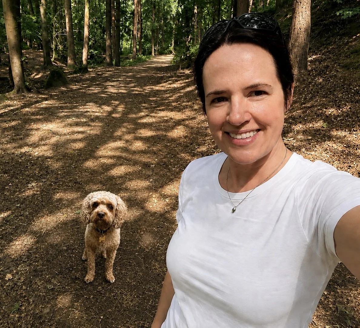 A woman taking a selfie in a wooded area with a dog sitting on a dirt path behind her.