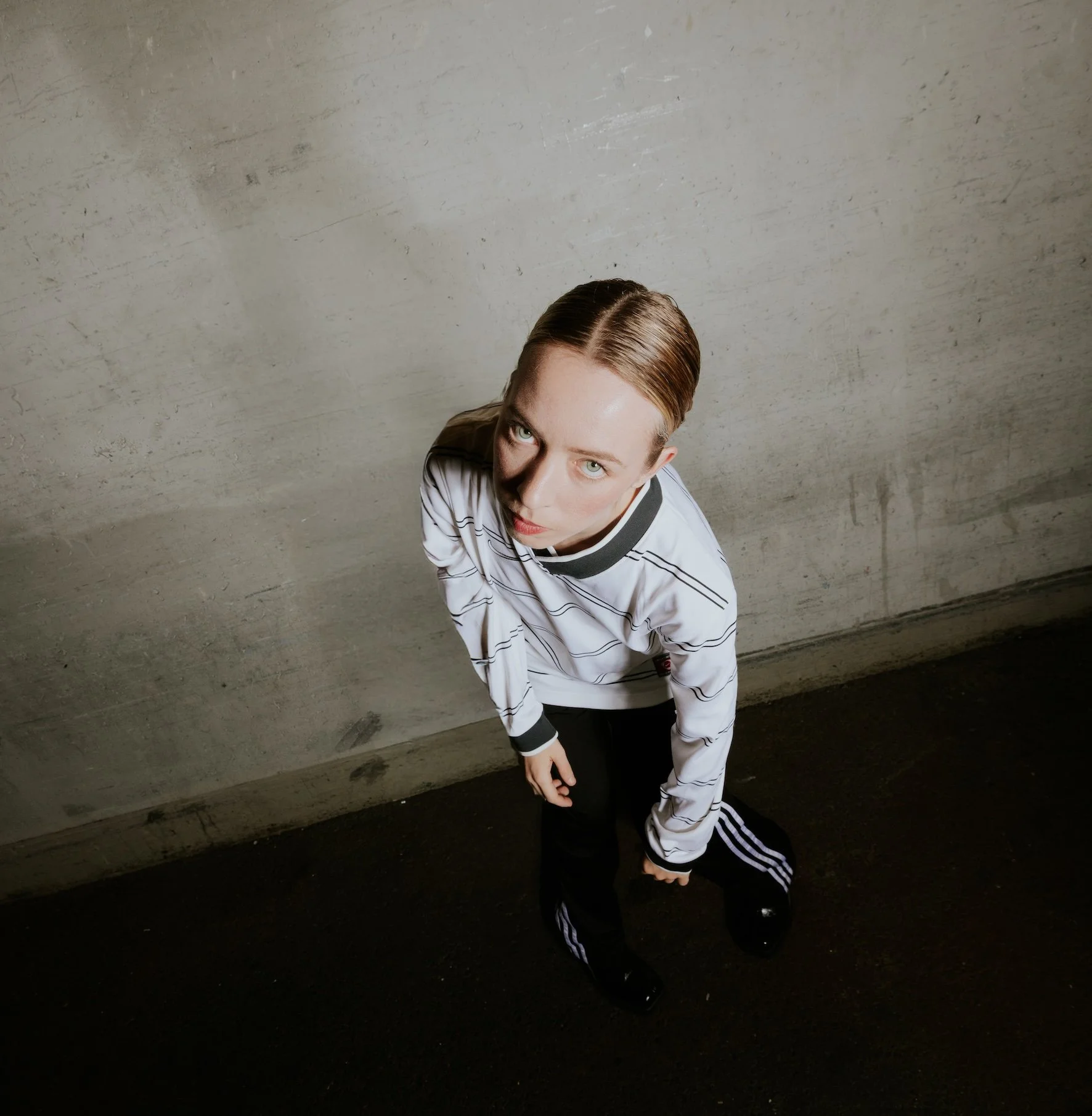 A woman with light skin and blonde hair, wearing a white and black striped sports jersey and black pants, kneeling and looking up at the camera against a concrete wall background.