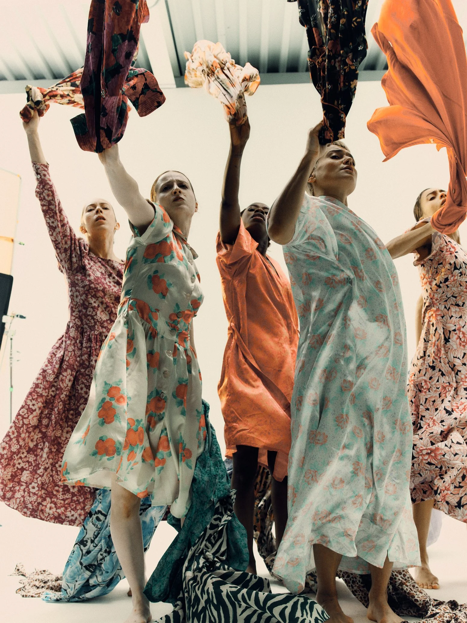 A group of people dressed in colorful, patterned clothing standing and kneeling in a studio with a plain white background, holding up garments above their heads.