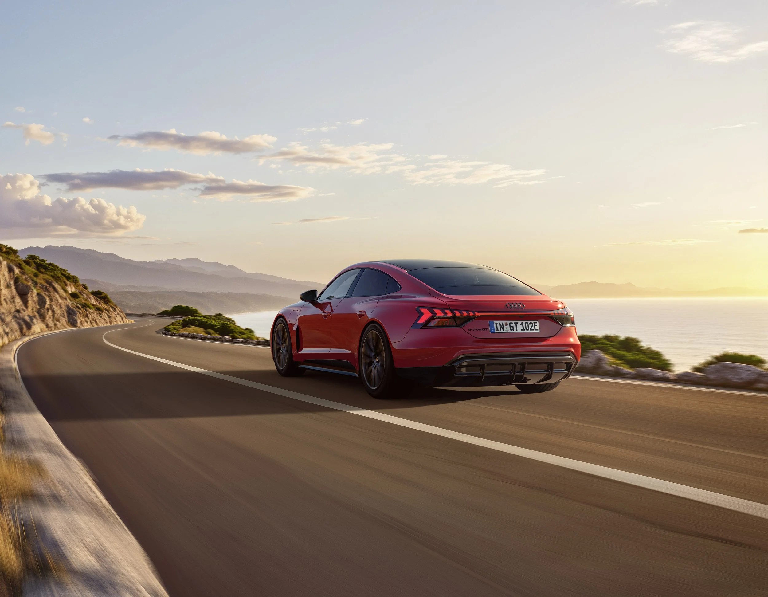 A red Audi e-tron GT driving on a winding coastal highway at sunset with mountains in the background.