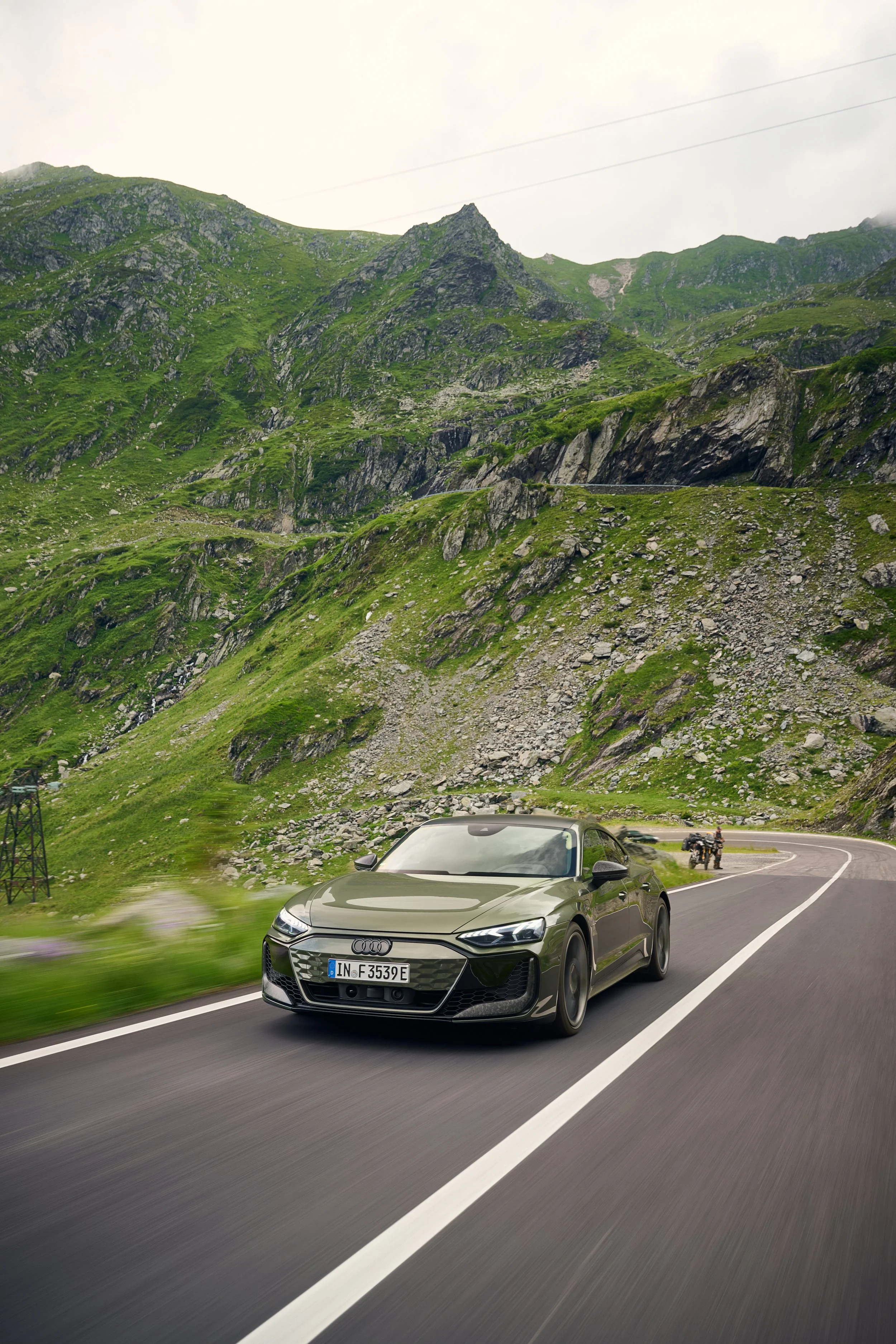 A gray Audi sedan driving on a winding mountain road with lush green hills and rocky terrain in the background, and a motorcyclist and another vehicle visible in the distance.