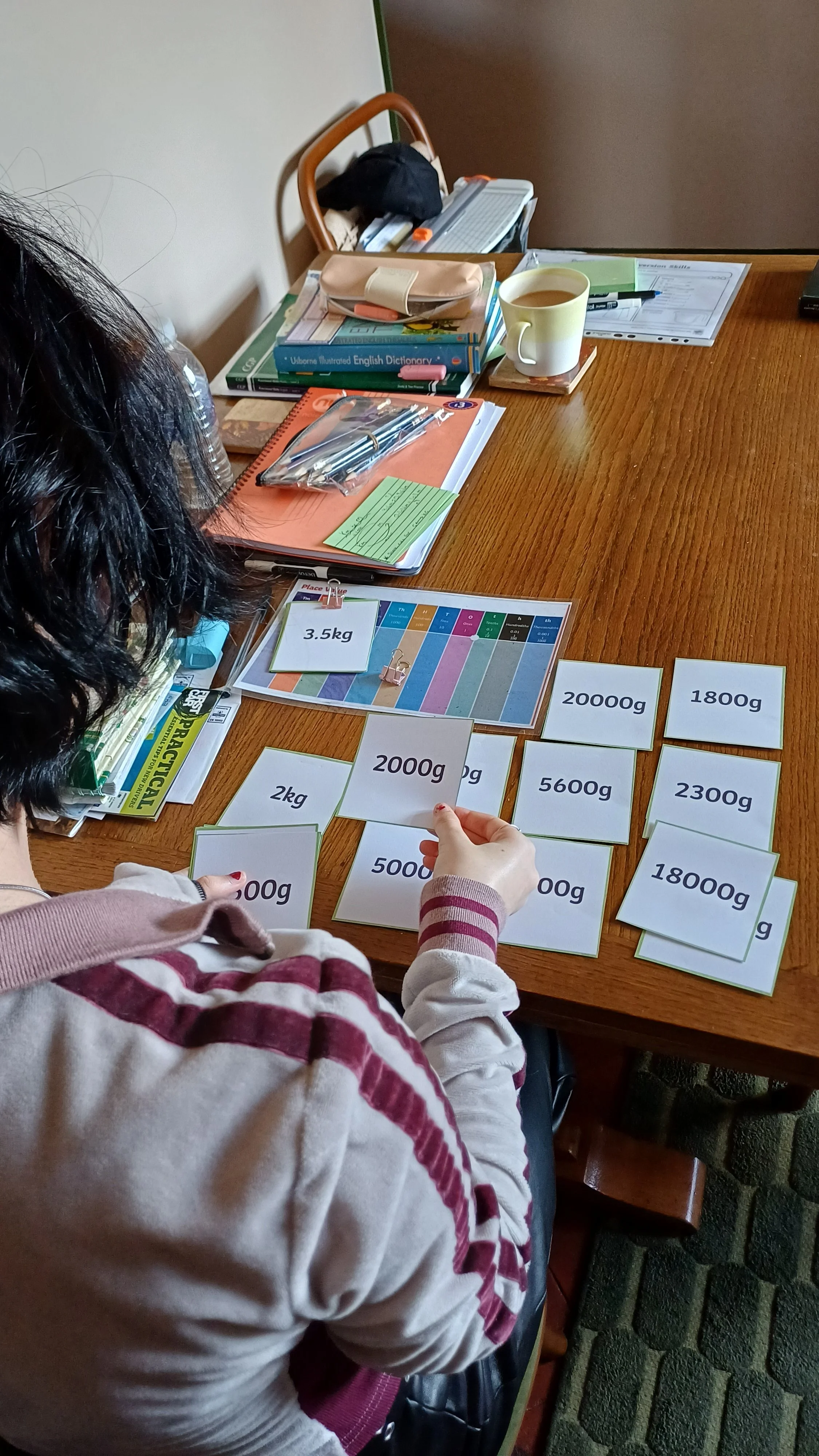 A person is organizing cards with weights written on them, such as 3.5kg, 2kg, 2000g, 5600g, 1800g, and 2300g, on a wooden table. The table also has notebooks, pens, a cup of tea, and various stationery.