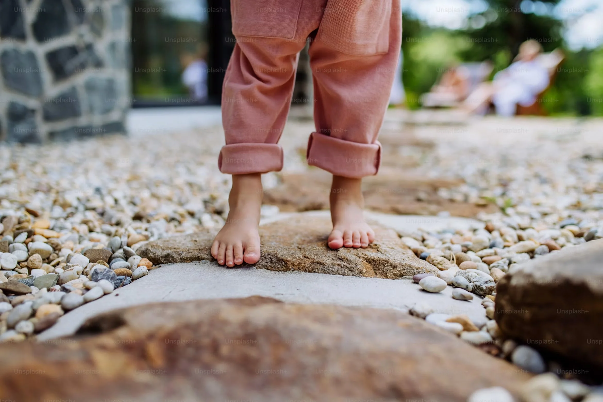 Close-up of a person's feet walking on stones along a beach, with their pink rolled-up pants and toes visible.