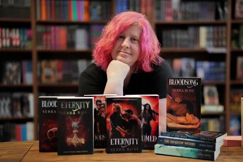 Serra Rose sitting at a table in a bookstore, surrounded by fantasy novel books, including titles like "Bloodsong," Consumed," & "Eternity."