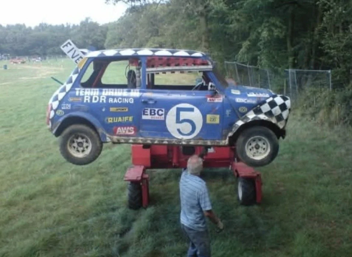 A race car, painted blue with various sponsor logos and the number 5, is elevated on a hydraulic lift in an outdoor grassy area. The car's wheels are off the ground, and a person stands underneath it, possibly inspecting or adjusting something.