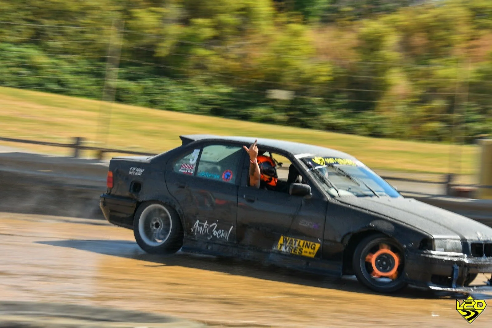 A black drift car on a dirt track, with a driver waving through the window, jokingly flipping the bird to the photographer , during a circuit.