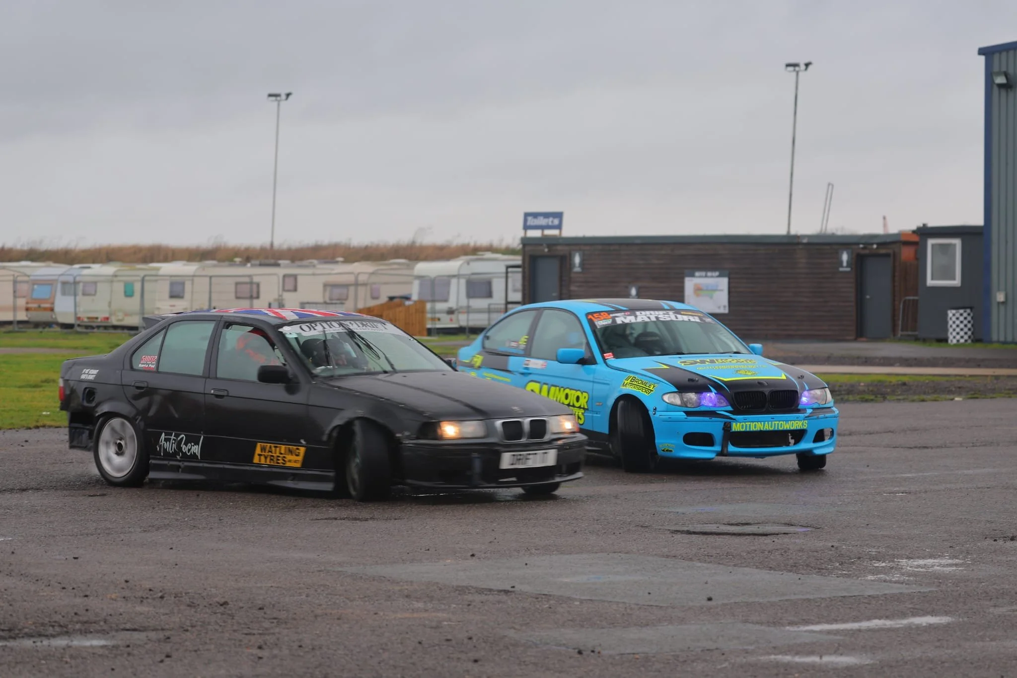 Two drift cars on a gray outdoor track, one black with yellow decal and the other blue with green decals, sliding side by side under a cloudy sky.