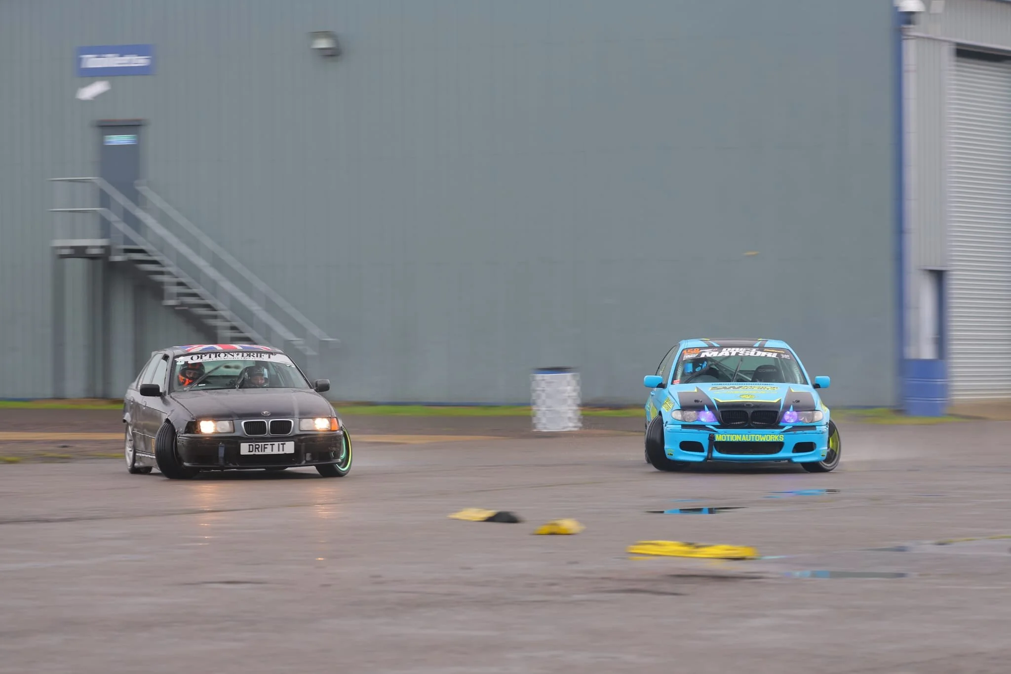 Two drift cars speeding on a track, one black with a license plate reading 'DRIFT IT' and the other blue with racing decals, with a large industrial building in the background.