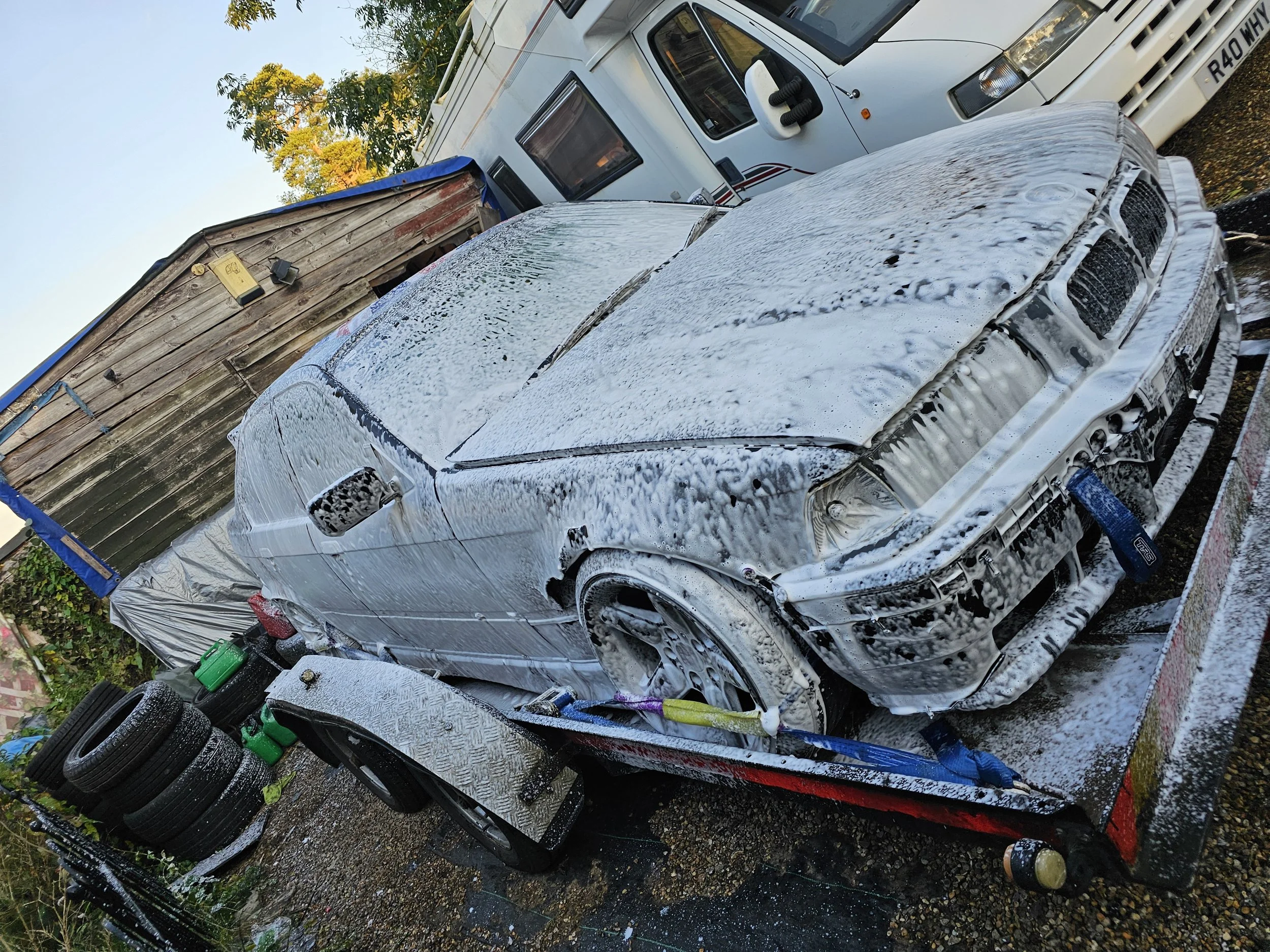A car covered in foam from a car wash, parked outside next to a wooden shed and a white camper trailer, with car tires stacked nearby.