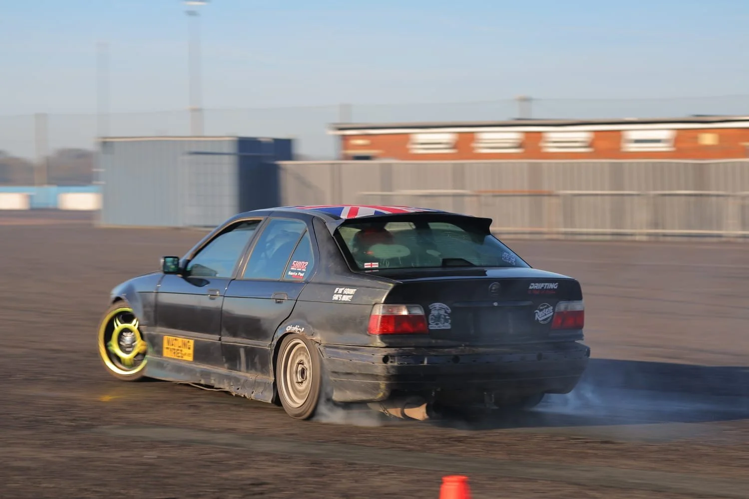 A black drift car with racing decals and a flag design on the roof, drifting on a dirt track, kicking up dust behind it.