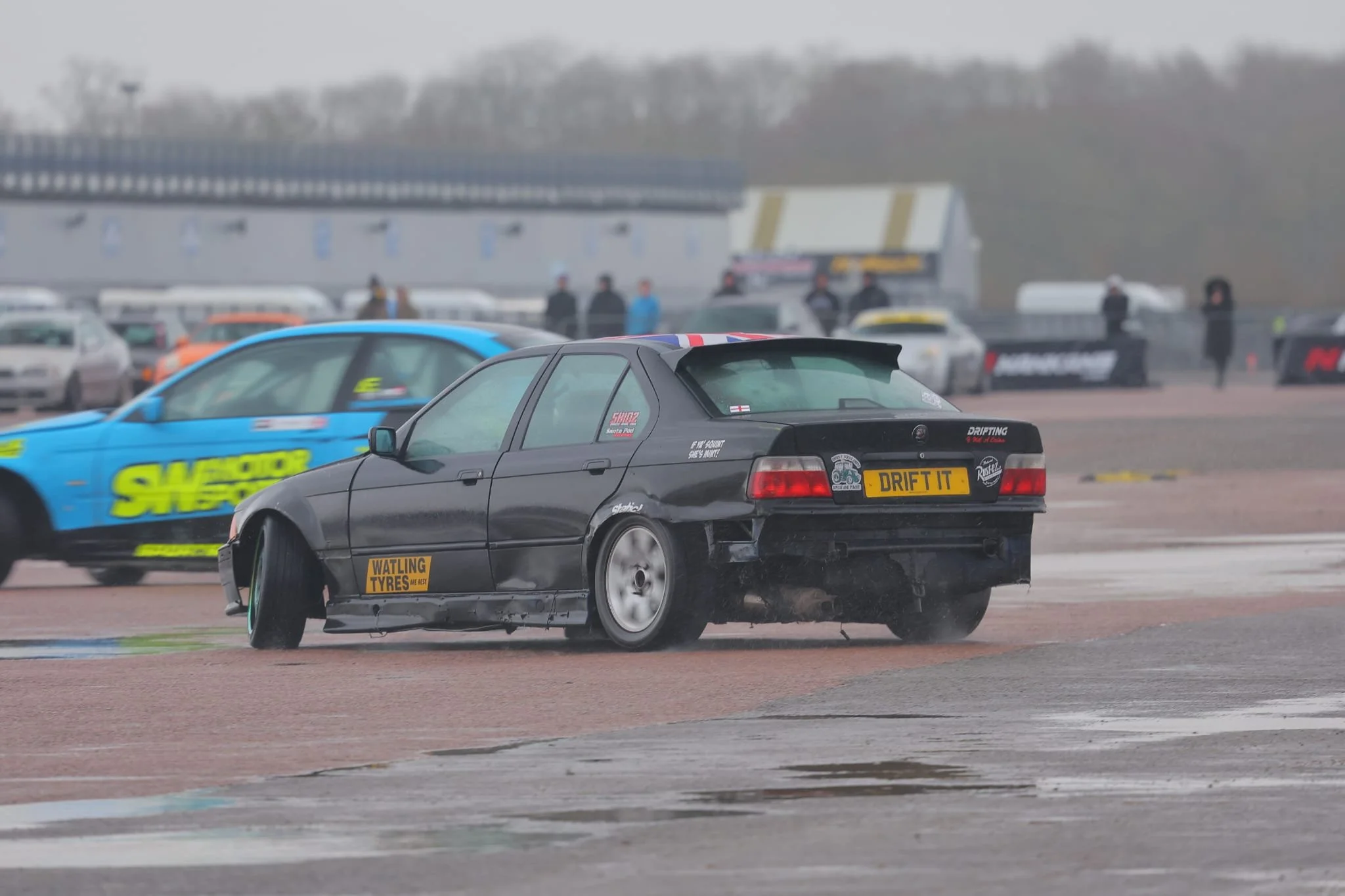 A black drift car with a yellow license plate that reads 'DRIFT IT' drifting on a race track, with a blue race car nearby, and several spectators and vehicles in the background on a cloudy day.
