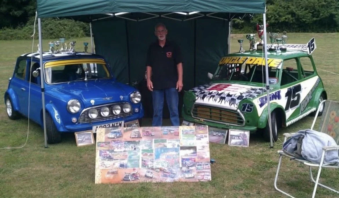 A man standing between two race cars under a green canopy at an outdoor event. The blue car on the left has multiple trophies displayed on top, and the green car on the right has racing decals and a wing with the number 15. Framed pictures and a coll