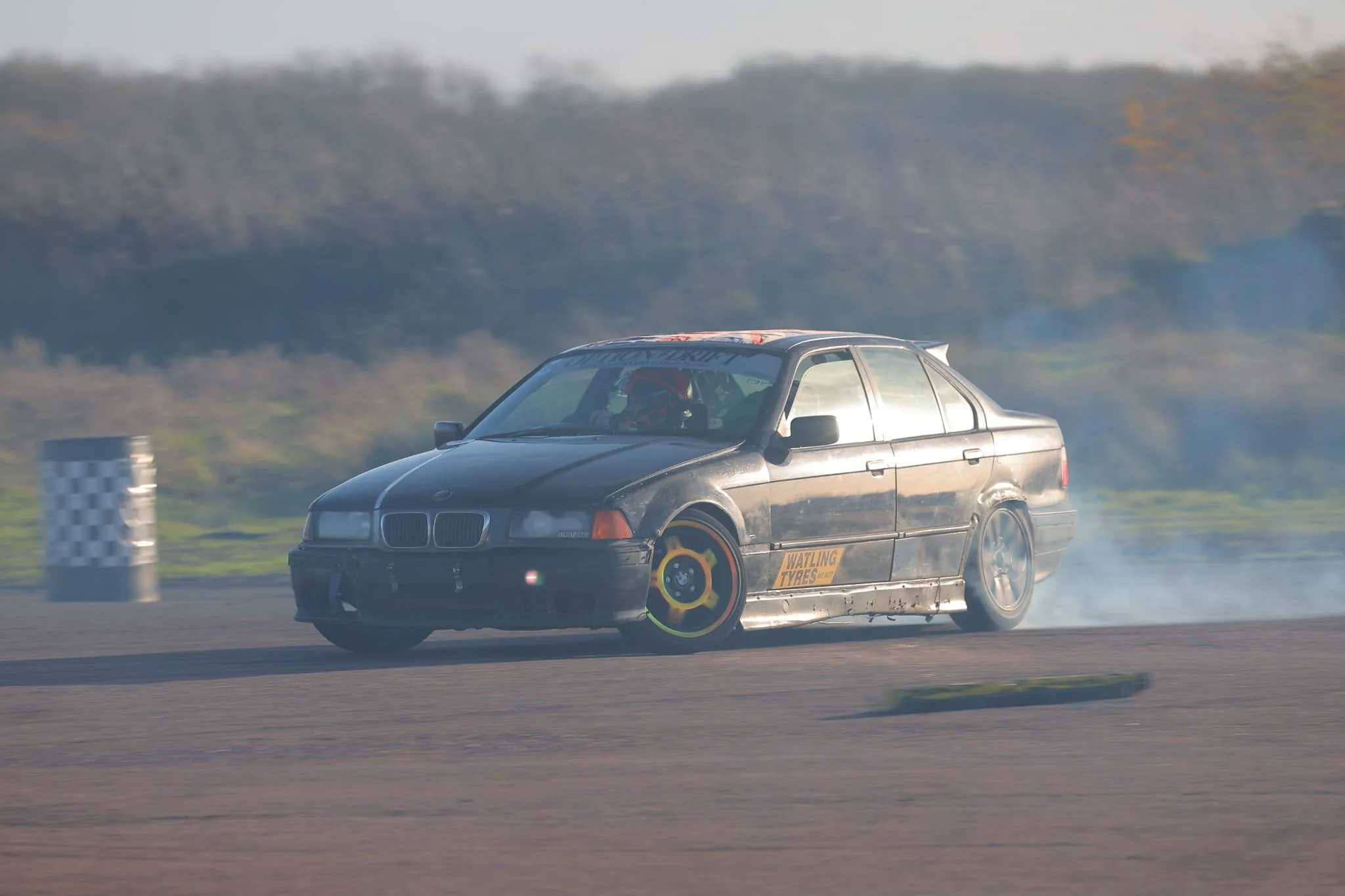 A black BMW car drifting on a race track with smoke coming from the rear tires, and a checkered flag in the background.