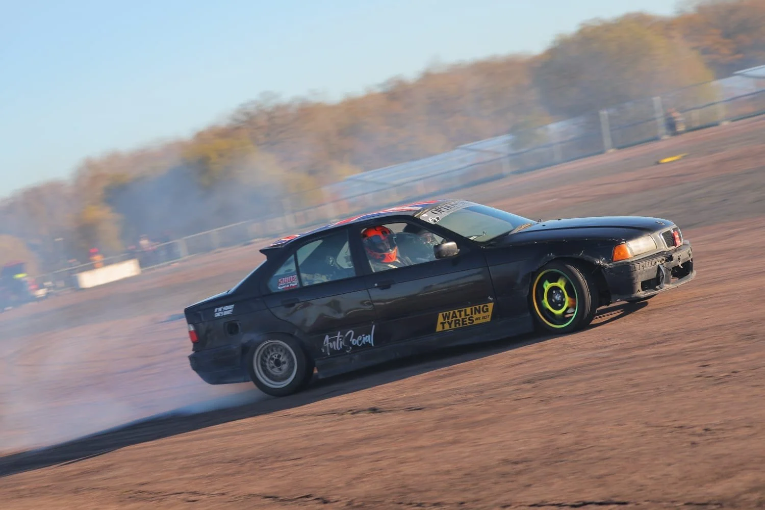 A black drift car drifting on a dirt track with tyres kicking up dust, driver wearing a helmet, autumn trees in the background.