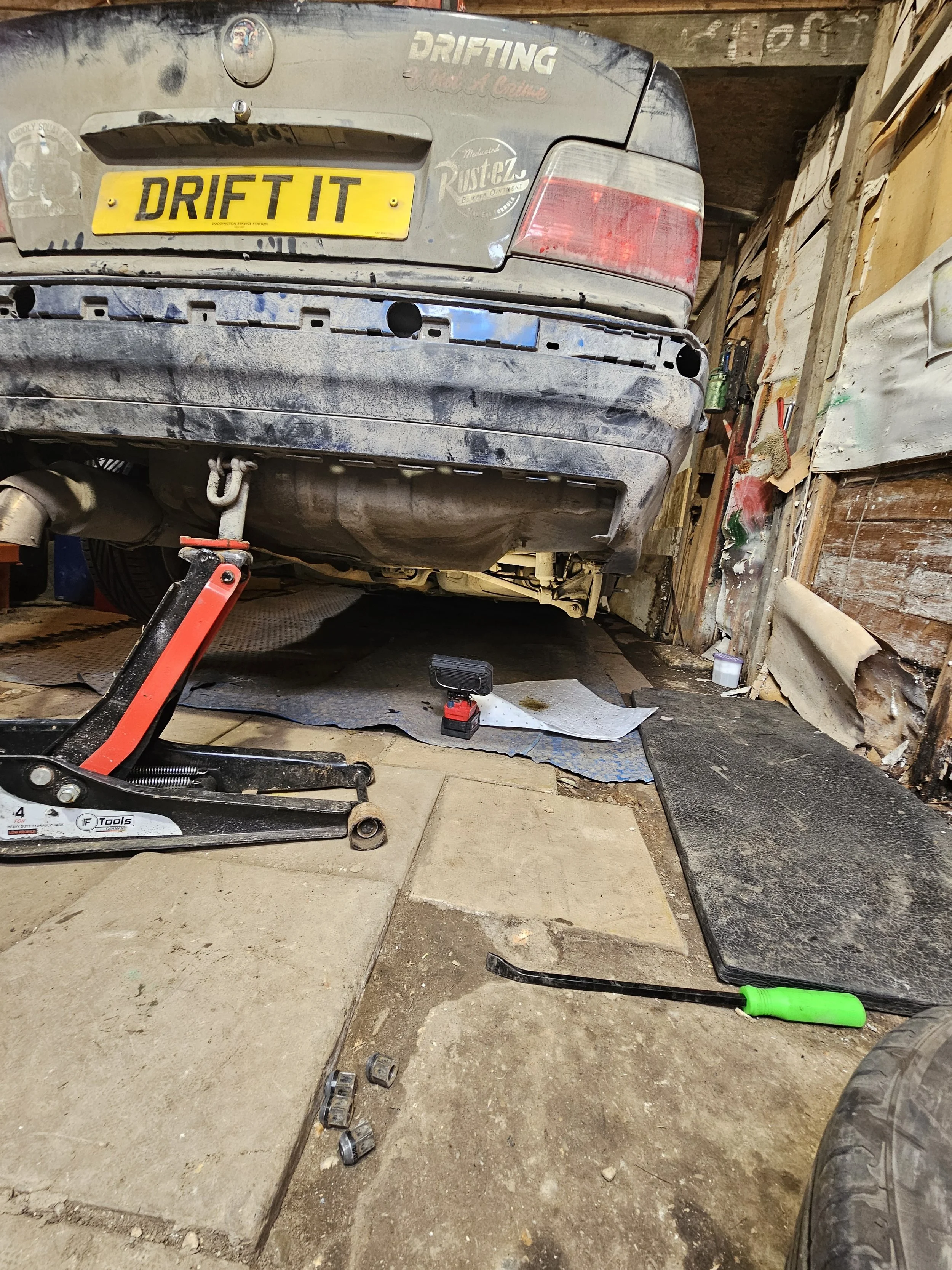 The image shows the rear view of a dusty, gray car in a workshop or garage, with a yellow license plate that says 'DRIFT IT.' The car's bumper is removed, and there are tools and car parts, including a hydraulic jack, scattered on the ground around i