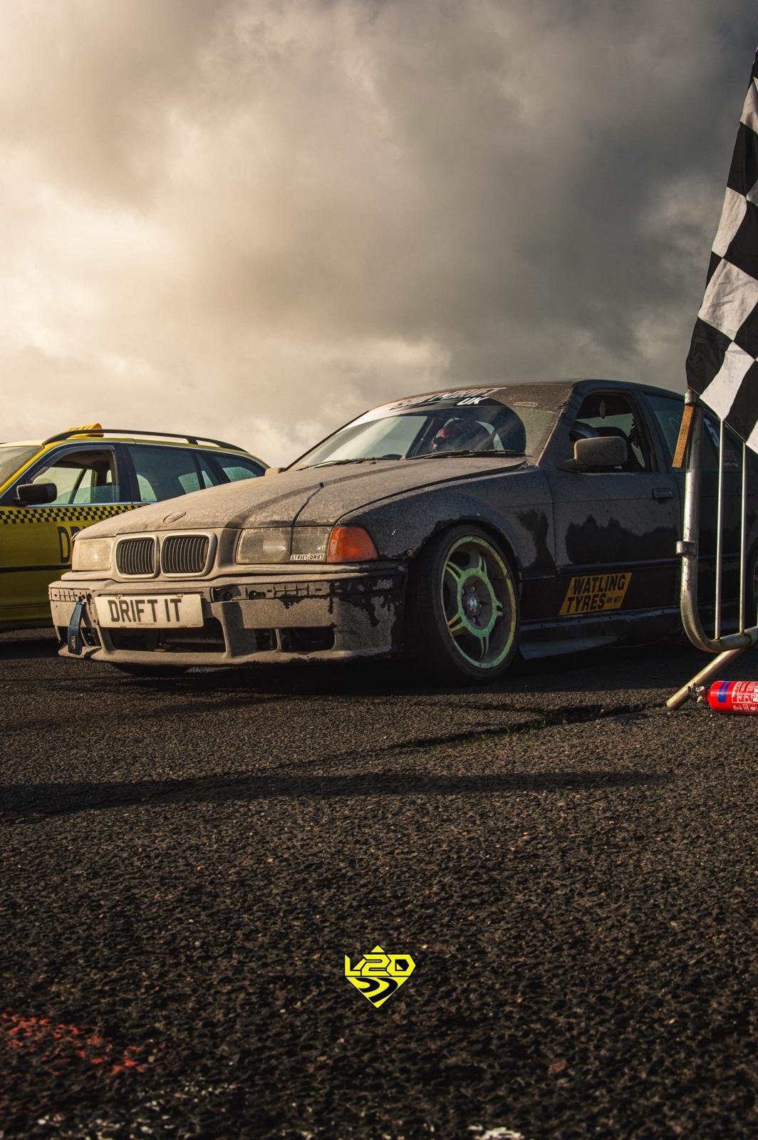 A black race car with a license plate that reads 'DRIFT IT', positioned next to a black and white checkered flag, with another vehicle and cloudy sky in the background.