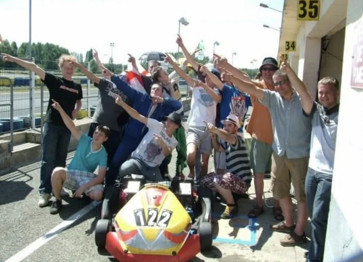 Group of people posing with a race car at a race track, all pointing upward and smiling.