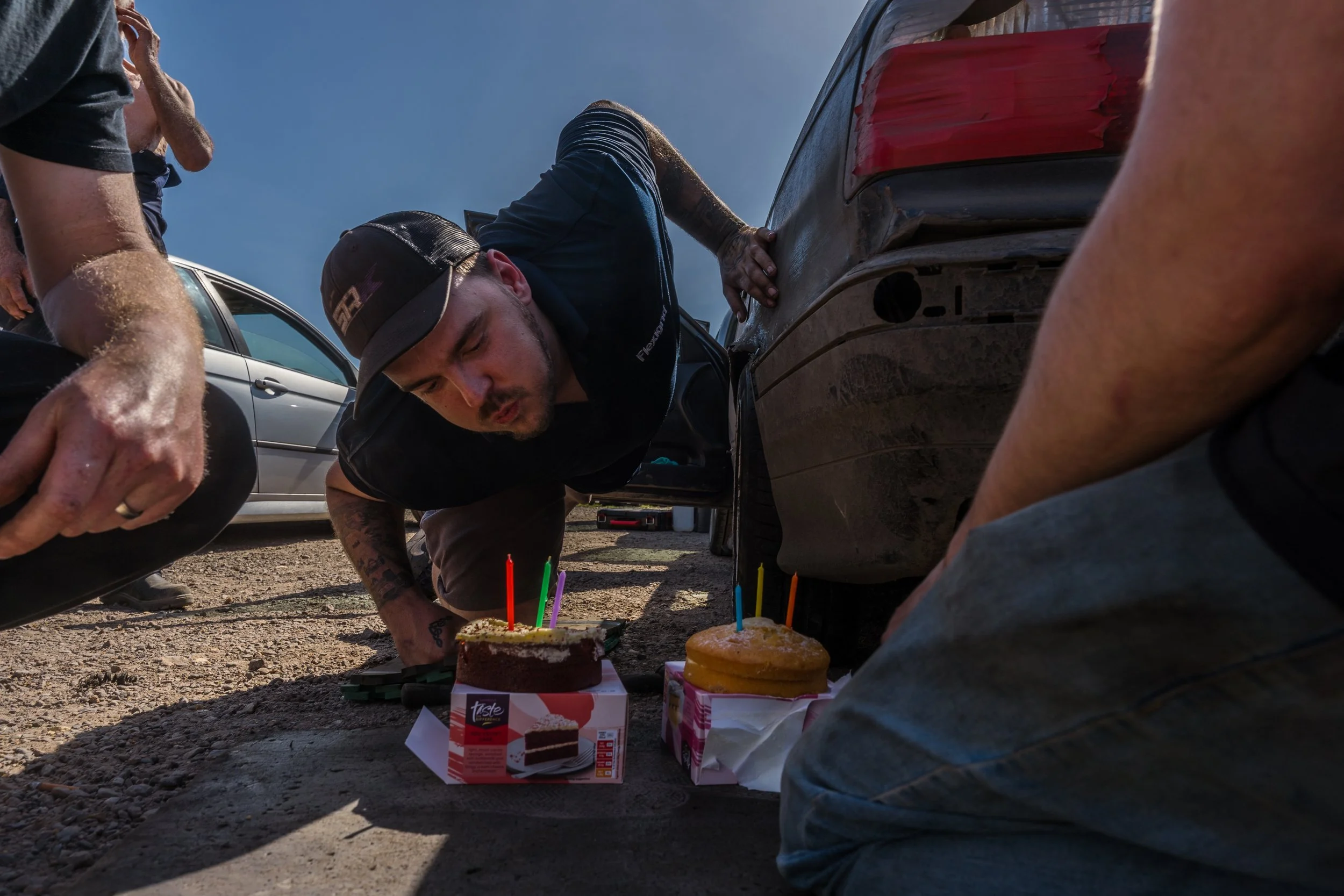 Multiple people gathered around celebrating clip point hires directors birthday whilst he kneels on the floor blowing the candles on his cake during a pit stop fix