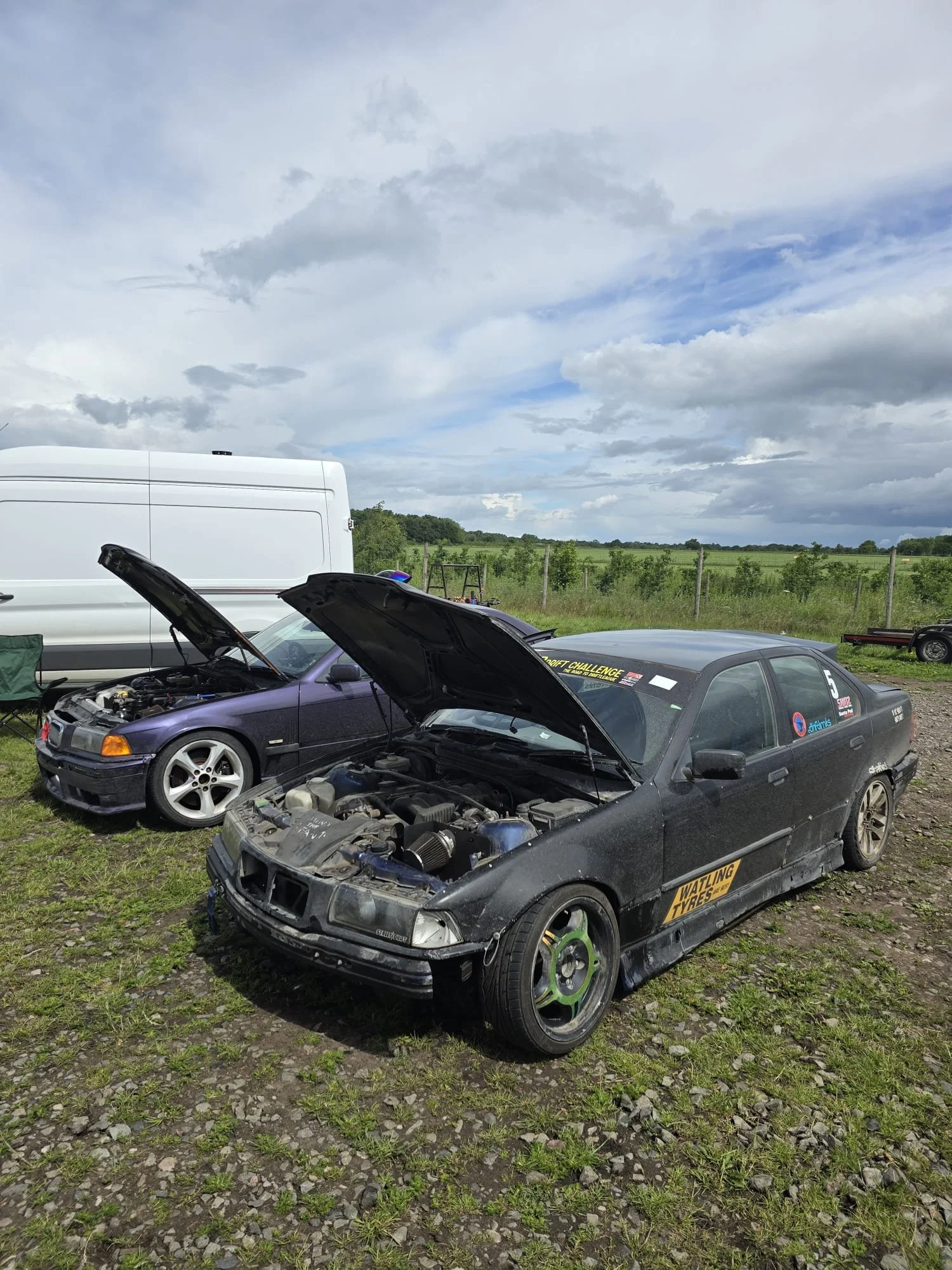 Three cars with open hoods parked on grassy ground, with a white van and a fenced field in the background under a cloudy sky.