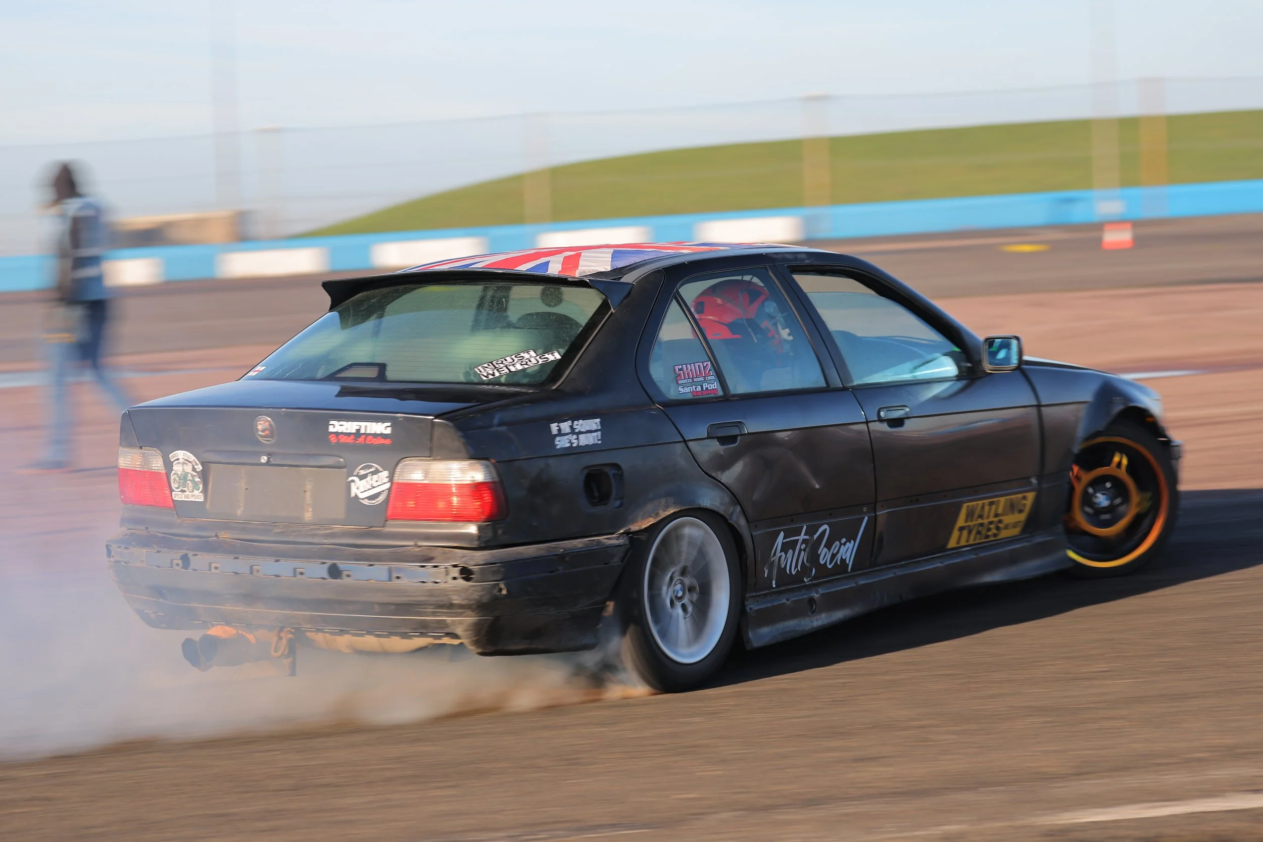 A black drift car with decals drifting on a race track, emitting smoke from the rear tires, with a blurred background indicating high speed.