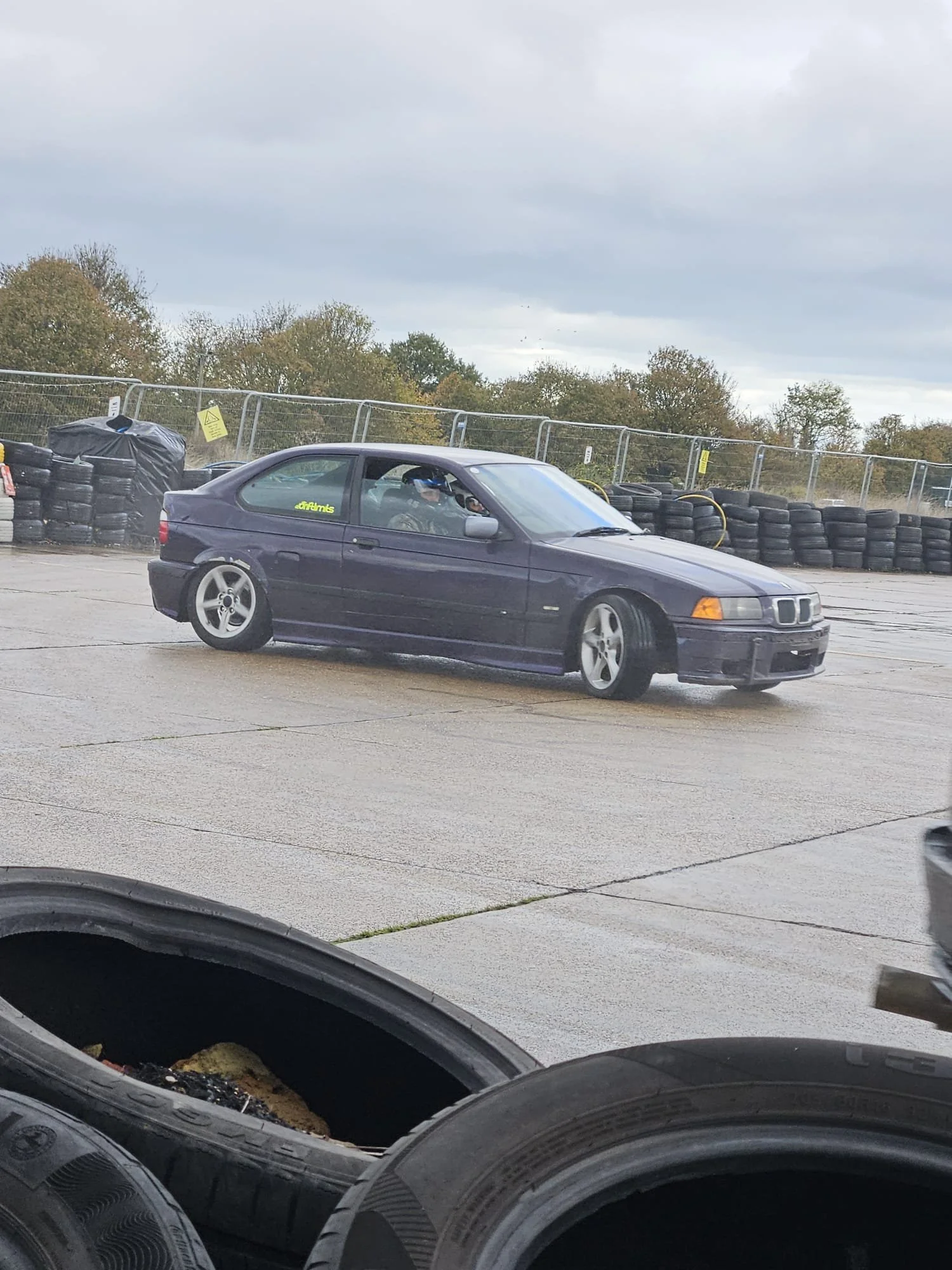 A blue BMW car on a race track with black tires and a driver wearing a helmet, surrounded by a safety fence and stacked tires, under a cloudy sky.