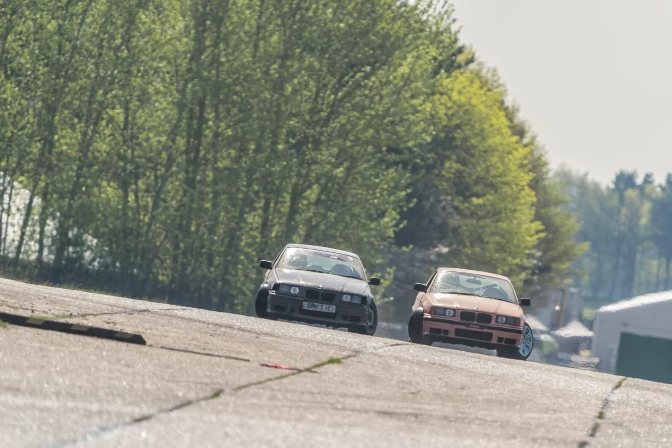 Two drift cars driven on a track with green trees in the background.