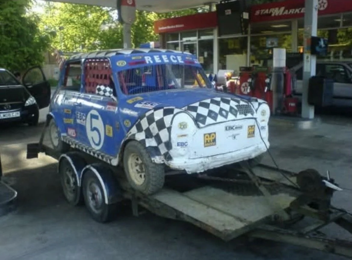 A vintage race car with a blue and white checkered design, labeled with the number 5 and the name 'REECE' on the windshield, is mounted on a flatbed trailer at a gas station.