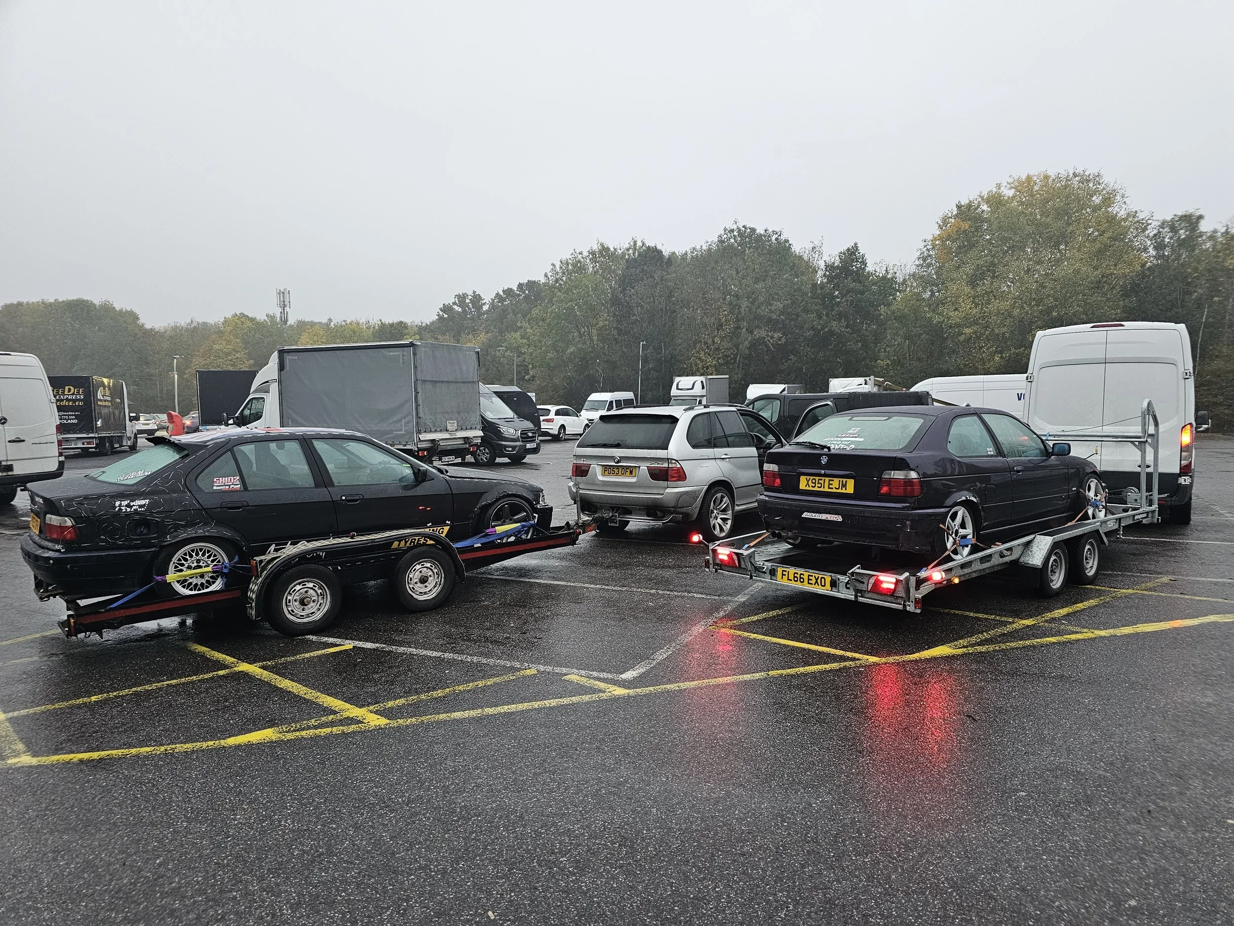 Overcast day in a parking lot with multiple cars and trucks, some on trailers, yellow parking space lines, trees in the background, and a cell tower visible in the distance.