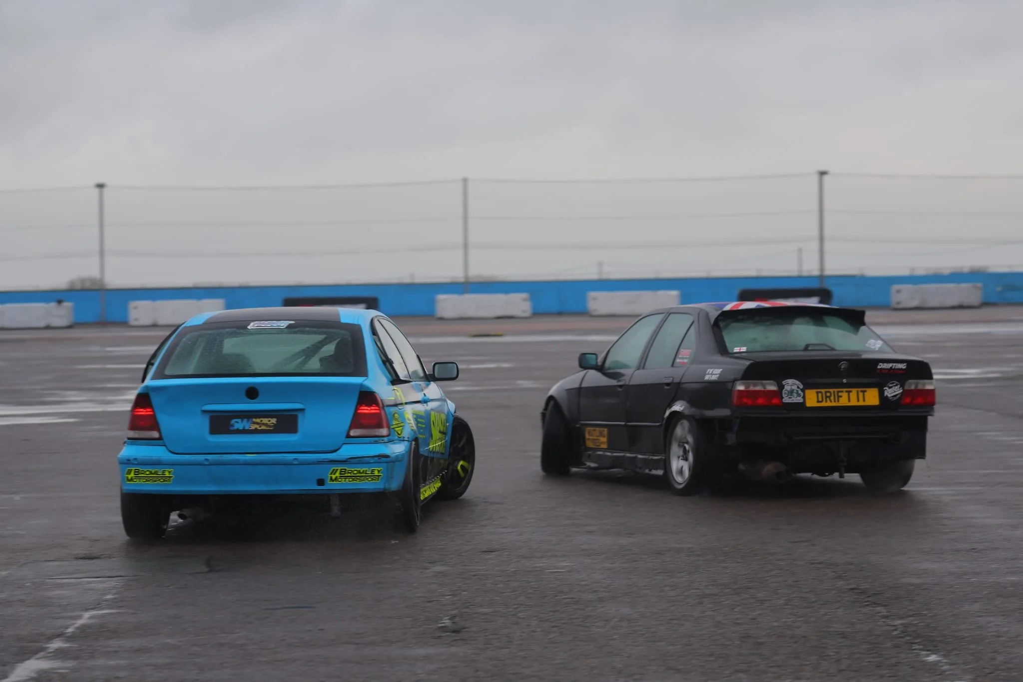 Two cars drifting on a track with a cloudy sky and barriers in the background.