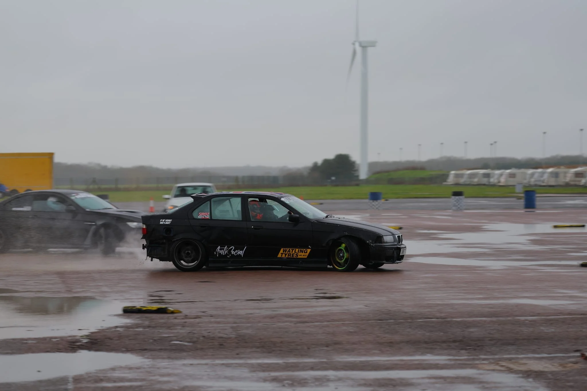 A black car with green rims is drifting on a wet track with water splashing behind it. There are other cars in the background, and a wind turbine is visible in the cloudy sky.