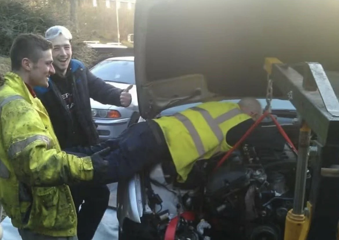 Clip point hires dad helping to fix an engine by being tipped upside down and held by his legs while he leans into the engine bay of a car