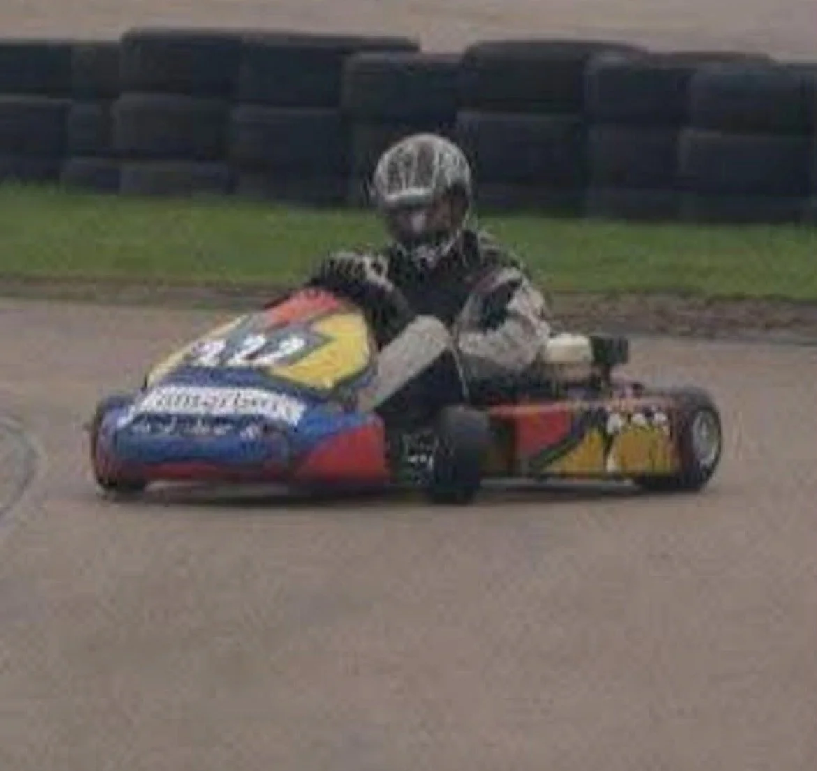 Clip point hire boss riding a go-kart on a race track, wearing a helmet and racing suit with a colorful design.