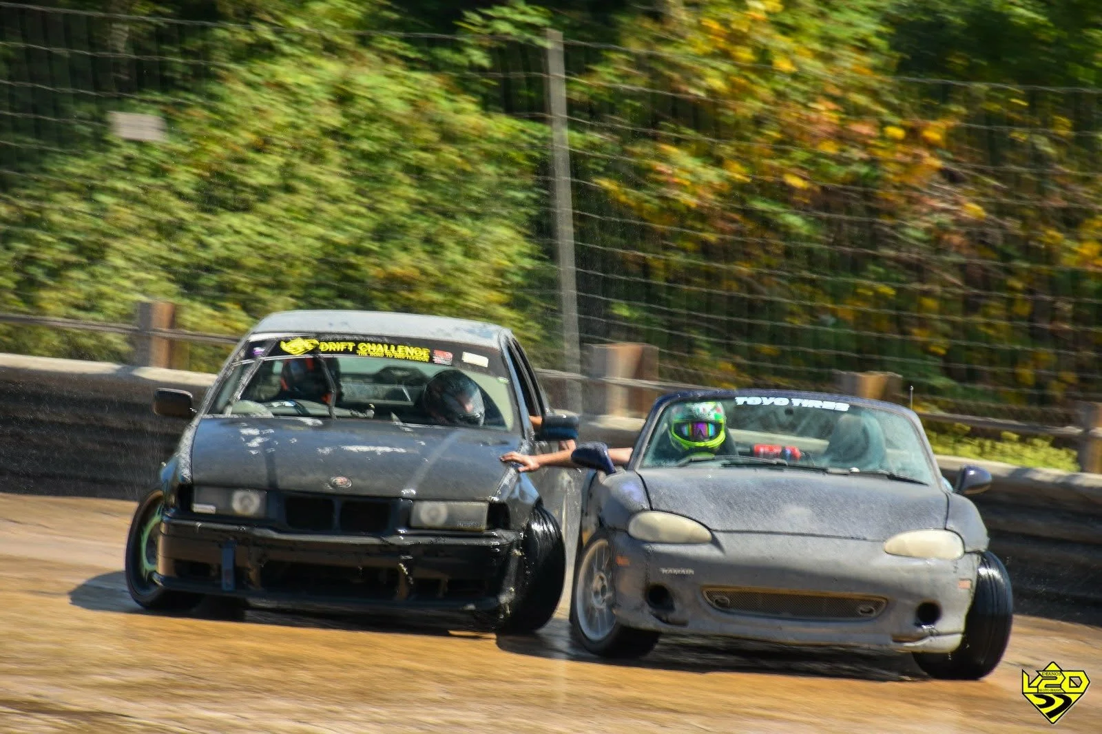 Two cars drifting next to each other so close that the driver of one and passenger of the other are shaking hands