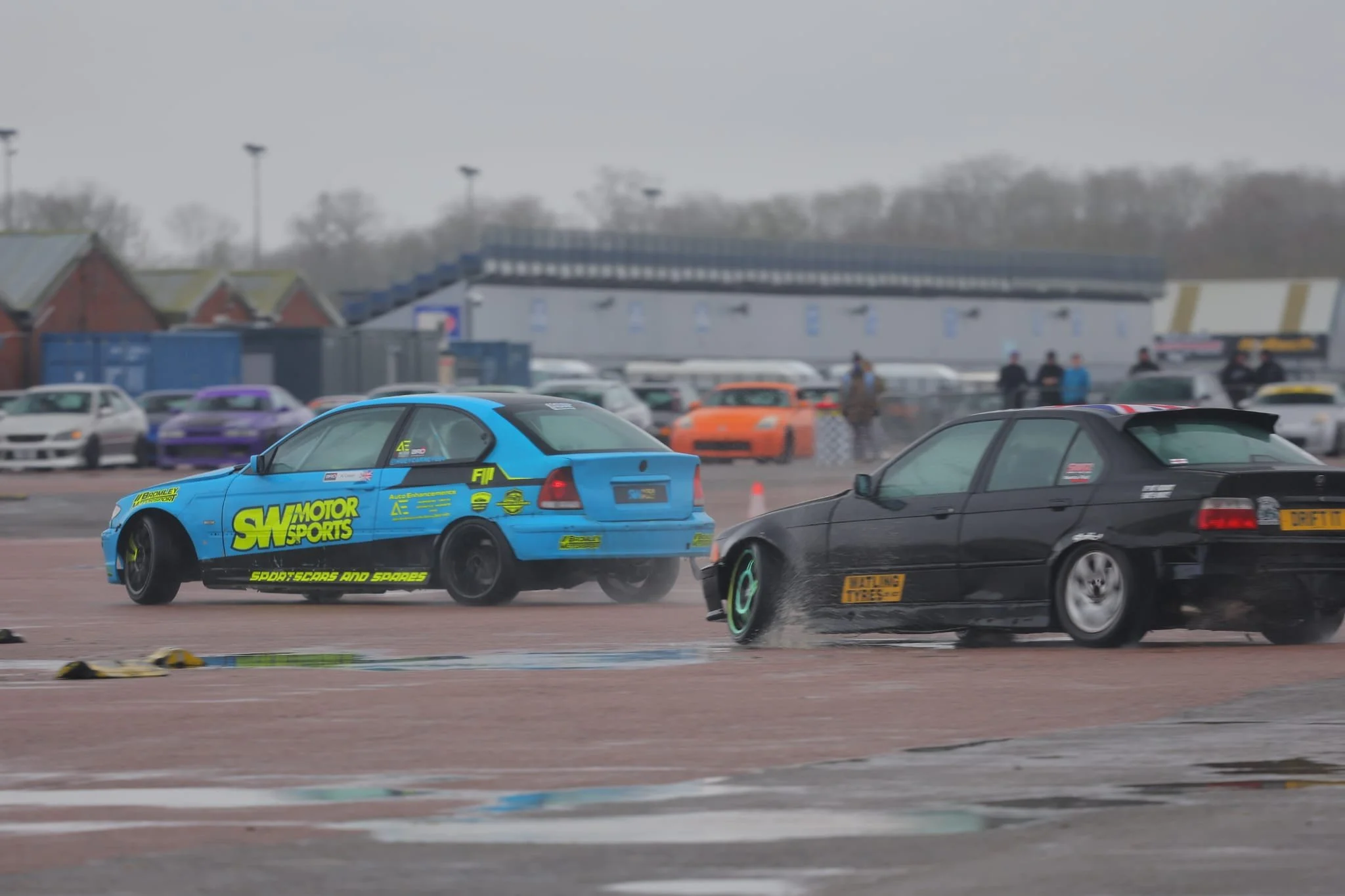 A blue drift car with yellow and black decals moving on a race track, with other cars and spectators in the background under an overcast sky.