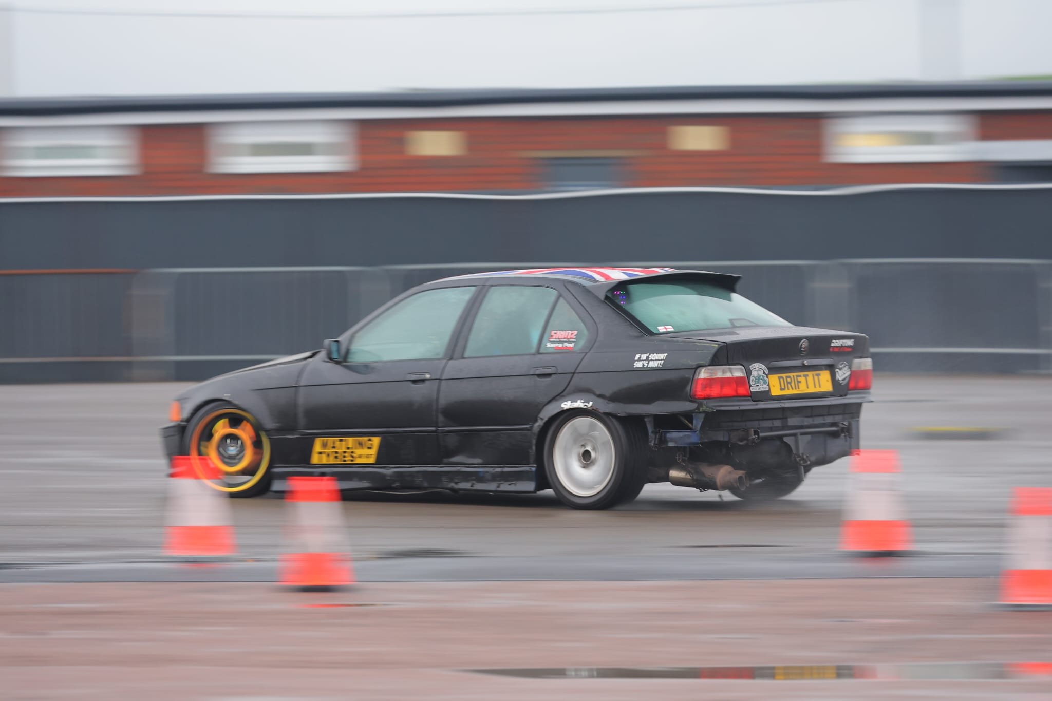 A black e36 car drifting around cones on a race track, with a Union Jack flag design on the car's roof and several stickers on the sides and rear, including one reading 'DRIFT IT!'