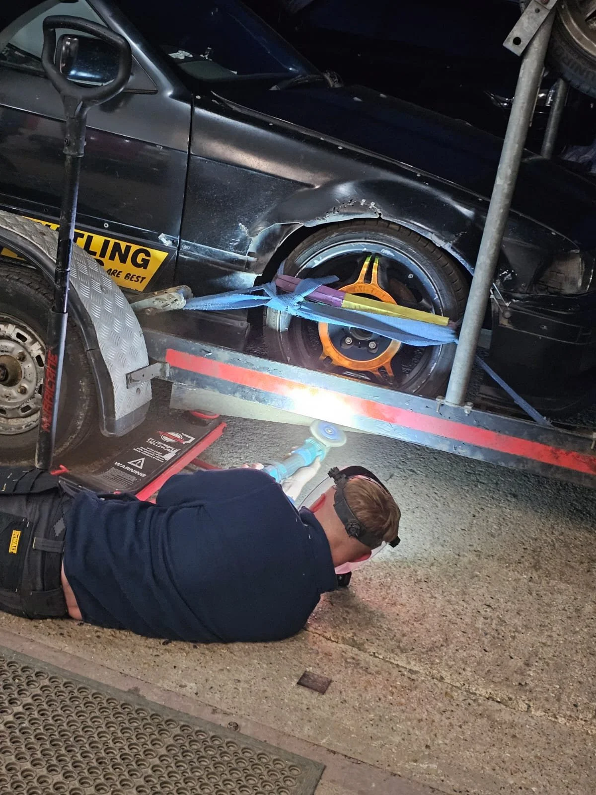 A man lying on the ground working on a black sports car that is secured on a tow truck. The car has a damaged front and a bright orange rim on the rear wheel. The man is using a power tool under the vehicle in a nighttime setting.
