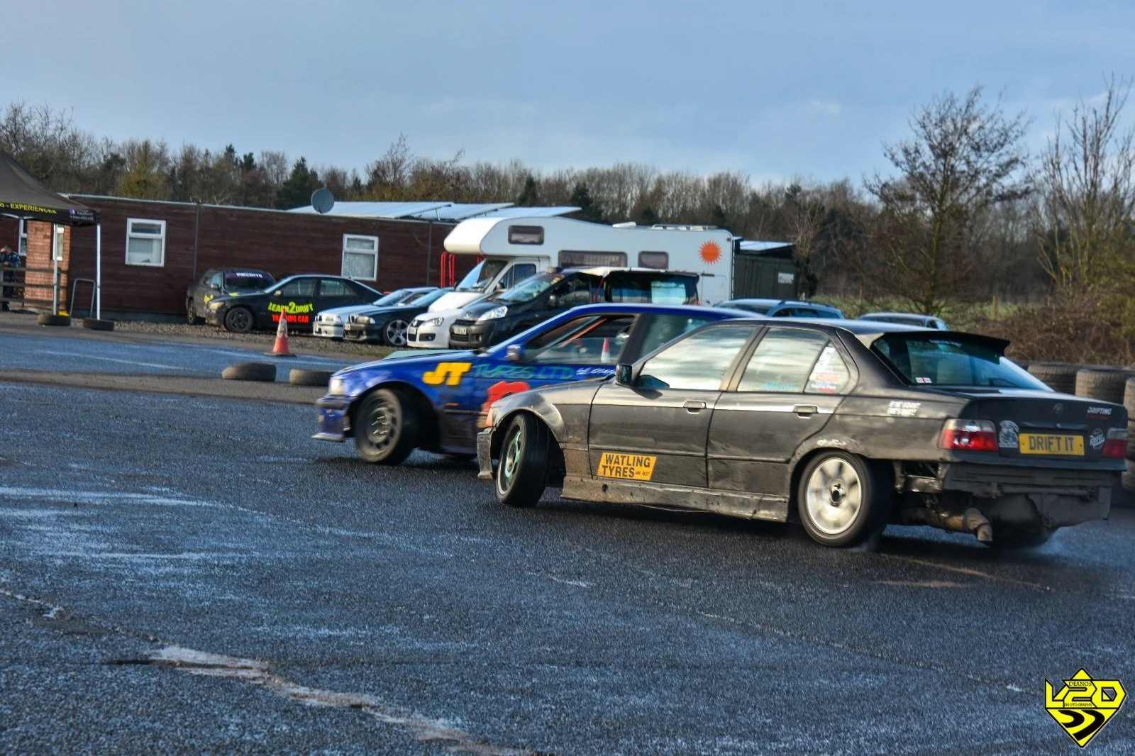 Two cars drifting on a race track, with multiple parked cars and a trailer in the background under a partly cloudy sky.