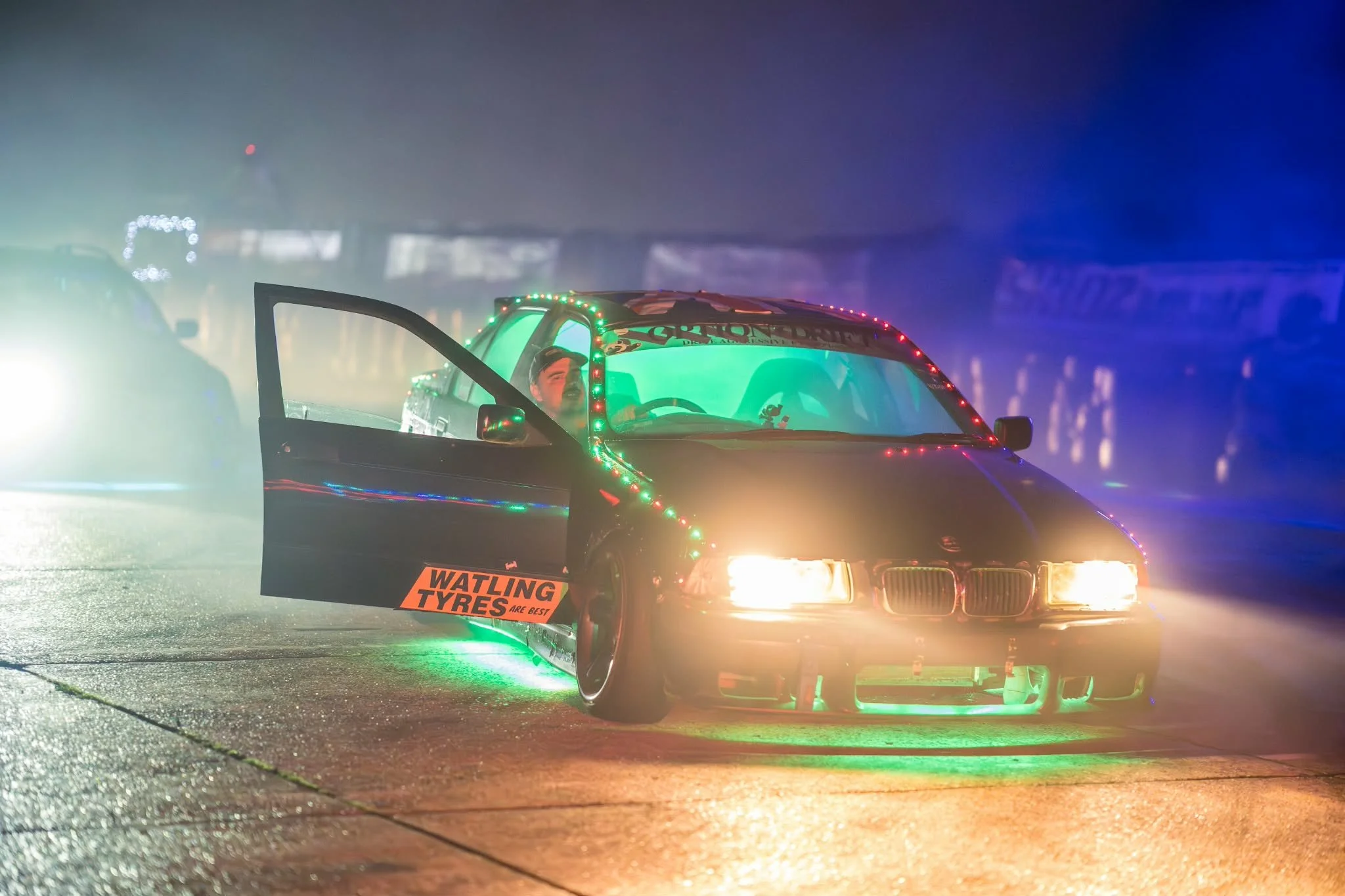 A race car decorated with colorful Christmas lights, sitting on a wet surface in a foggy or misty environment.