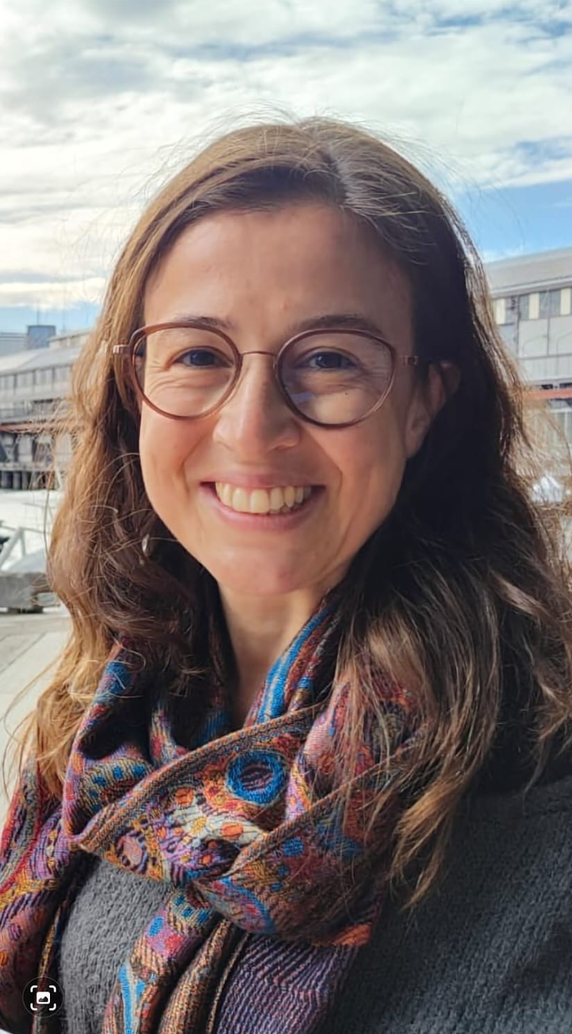 A smiling woman with long brown hair, glasses, and a colorful scarf outdoors with a cloudy sky and modern buildings in the background.