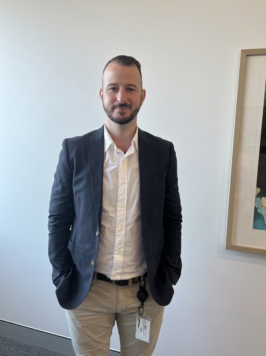 A man with short dark hair, a goatee, wearing a white shirt, navy blazer, and khaki pants, standing indoors against a plain white wall with a framed picture on the right.