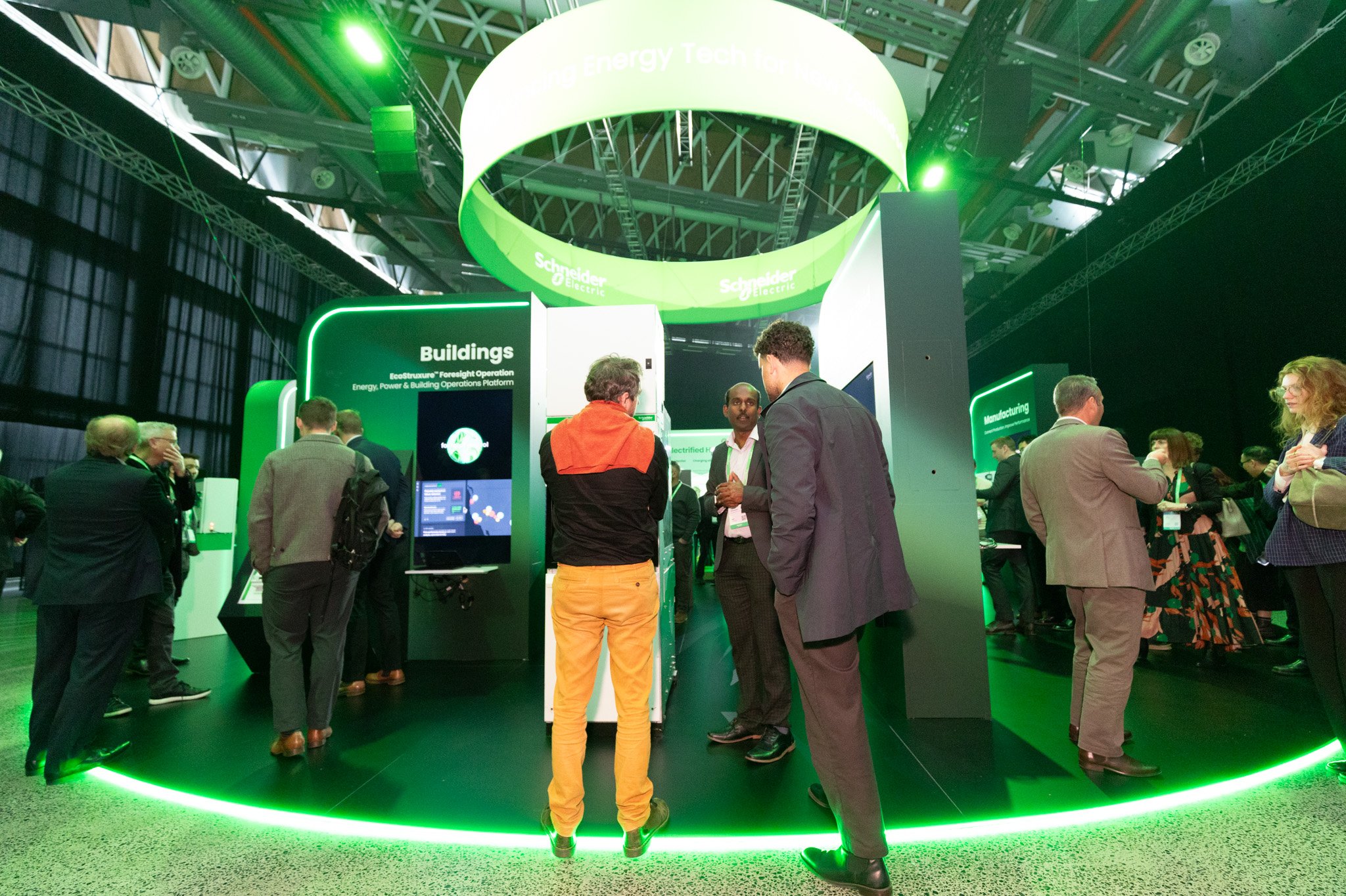 People attending a trade show or conference at a booth with green lighting and branding for Schneider Electric. The booth has informational displays and a large circular overhead sign reading 'Buildings' and 'Energy Technology.'