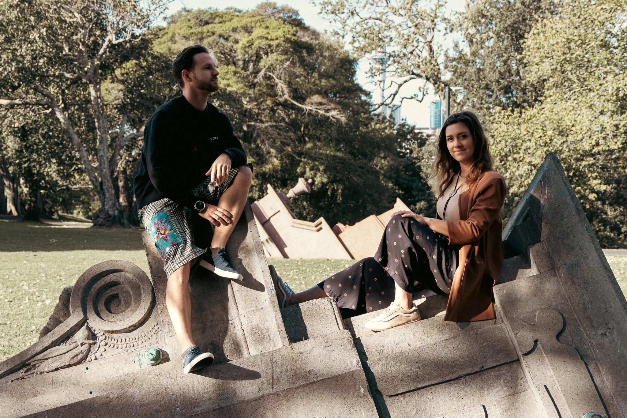 Two young adults, a man and a woman, sitting on a carved stone sculpture outdoors surrounded by trees. The man is sitting on the sculpture with one leg bent, wearing a black shirt and checkered shorts with floral print, while the woman is sitting on 
