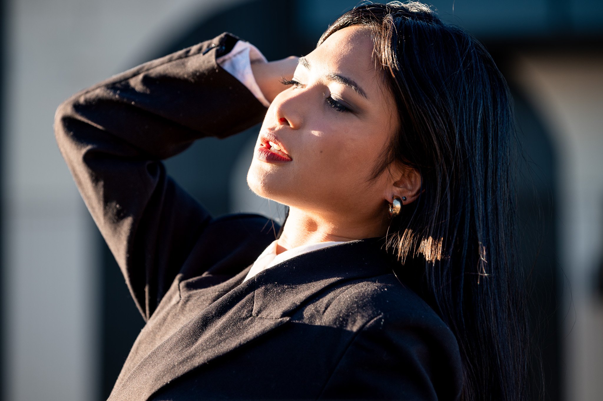 A close-up portrait of a woman with short dark hair, wearing earrings and a black jacket, with her eyes closed and her hand resting on her head, outdoors in sunlight.