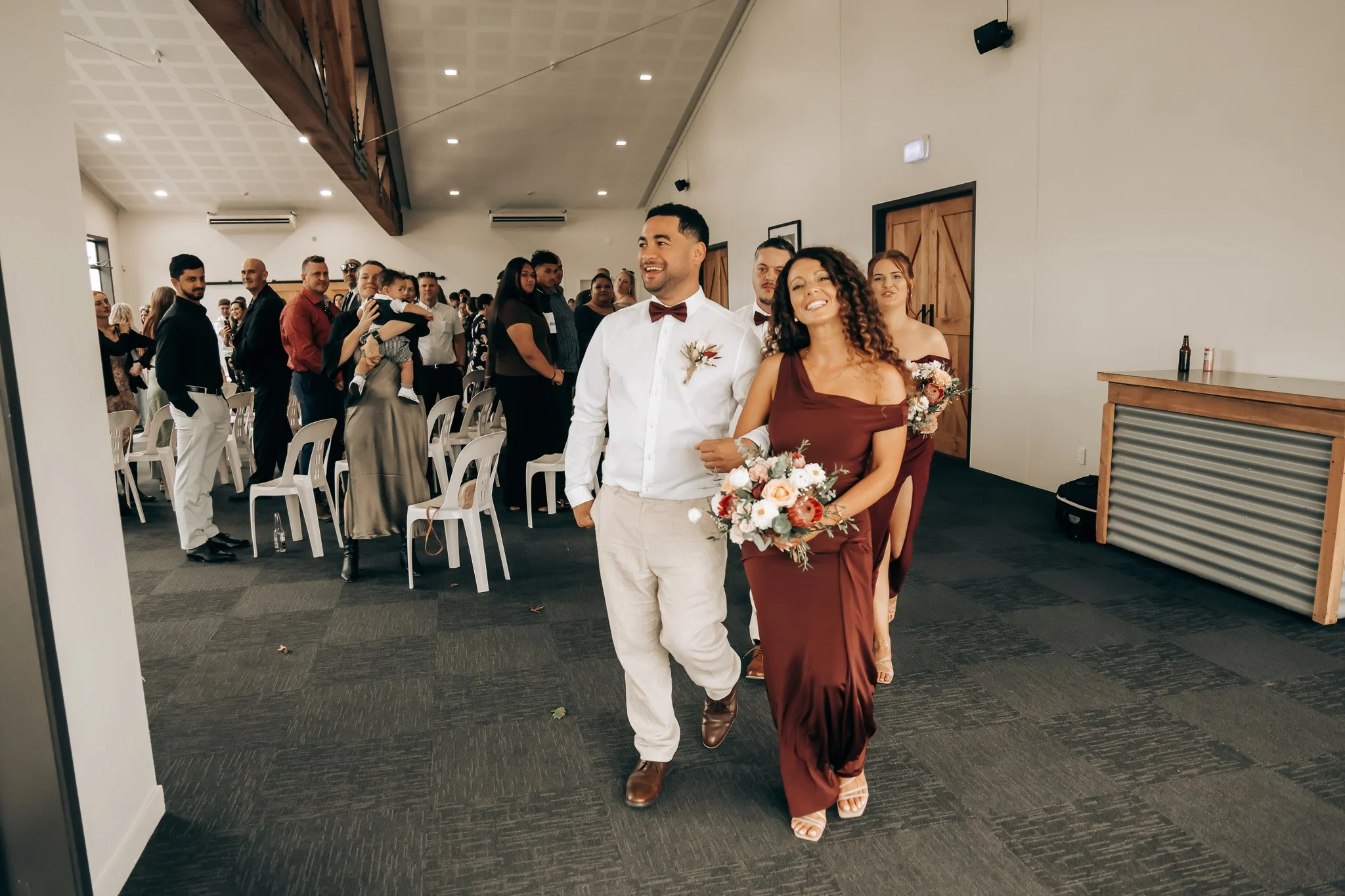 A wedding procession with a man in a white shirt and red bow tie, and a woman in a burgundy dress holding a bouquet, walking through a large room filled with seated guests.
