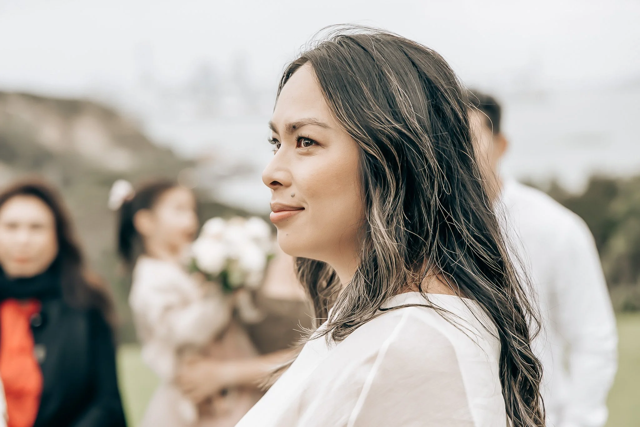 A woman with long, wavy hair and a small beauty mark on her face, smiling slightly, at an outdoor gathering with blurred people in the background.