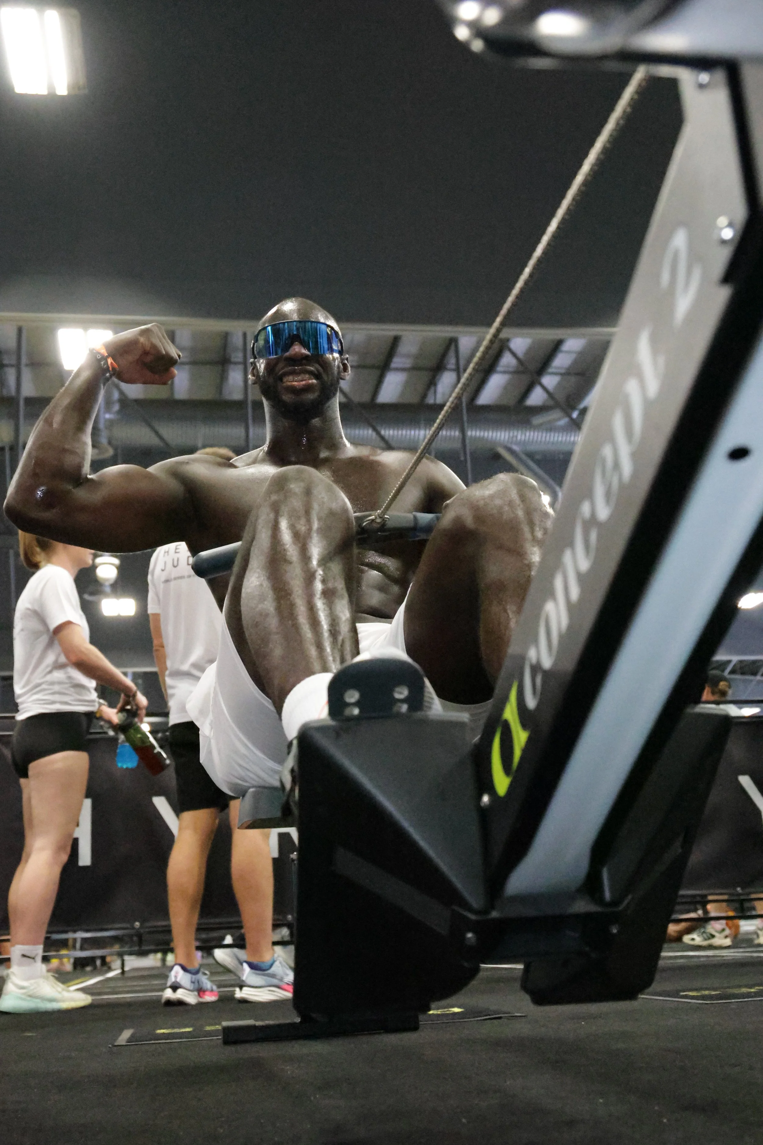 A muscular man wearing sunglasses flexes his bicep while sitting on a rowing machine at a gym, with other people in the background.