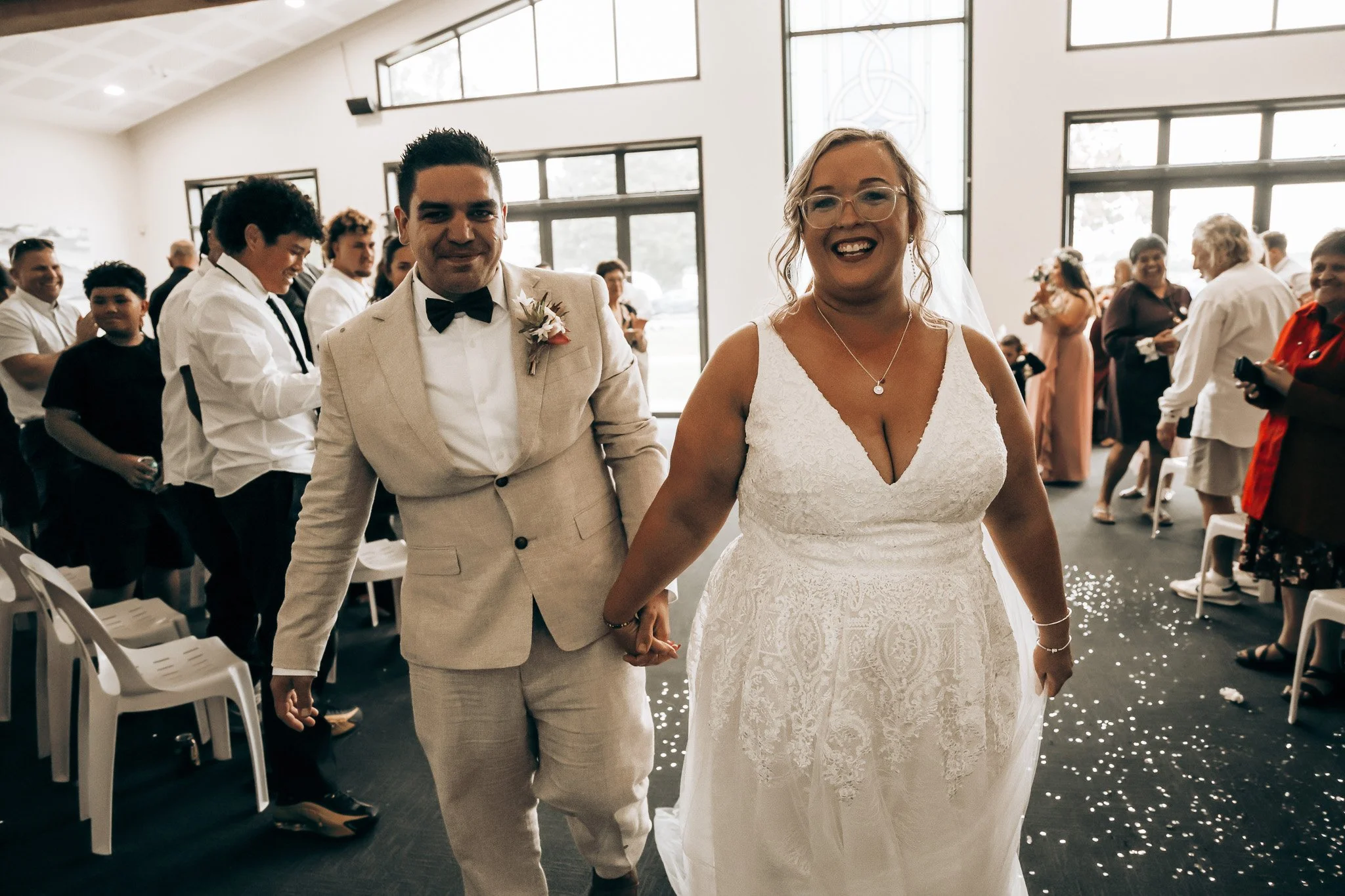 A bride and groom holding hands and smiling at a wedding ceremony inside a bright room with large windows and wedding guests in the background.